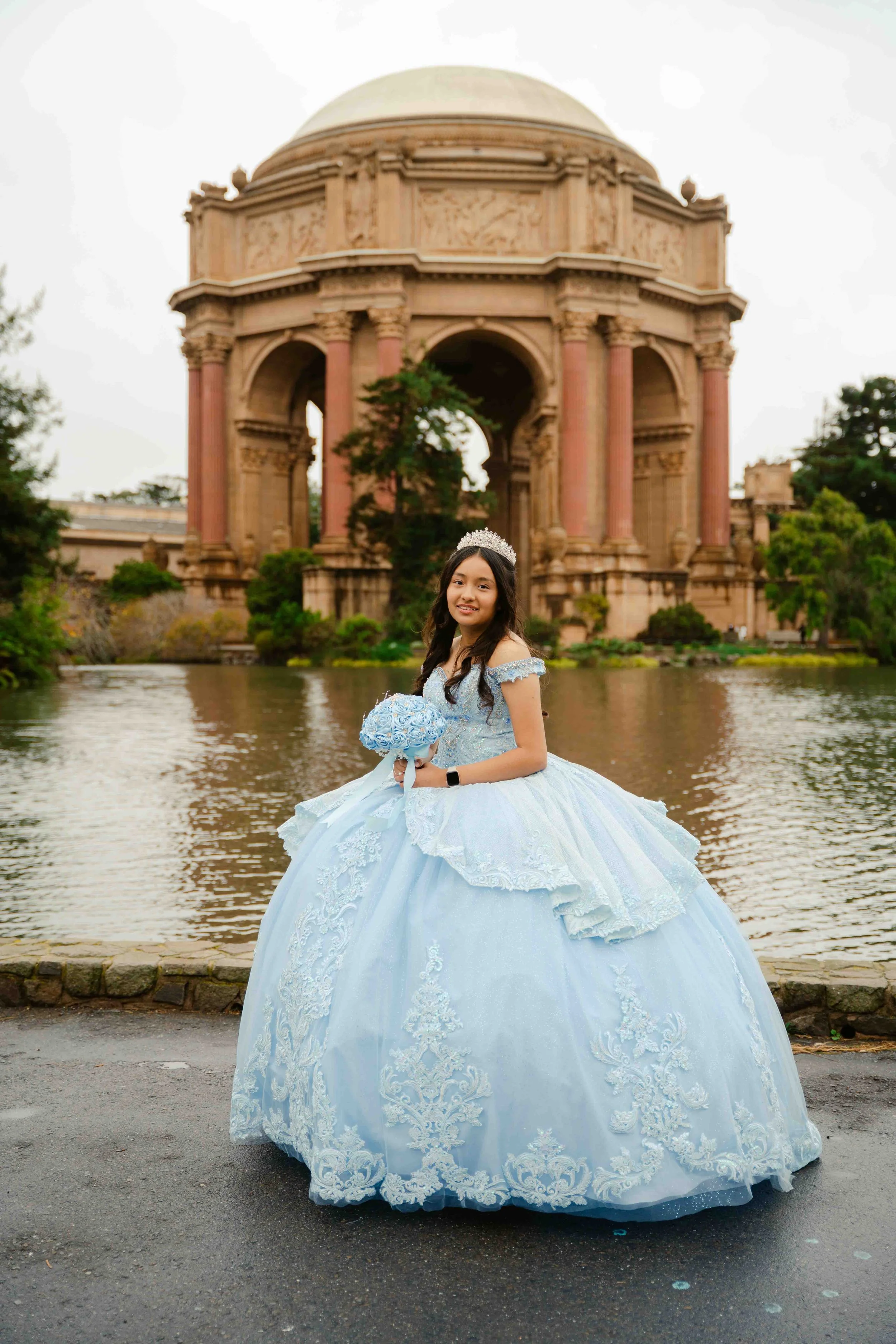 Young girl in a light blue quinceañera dress, wearing a tiara, holding a bouquet, standing near a lake with a historic building in the background.