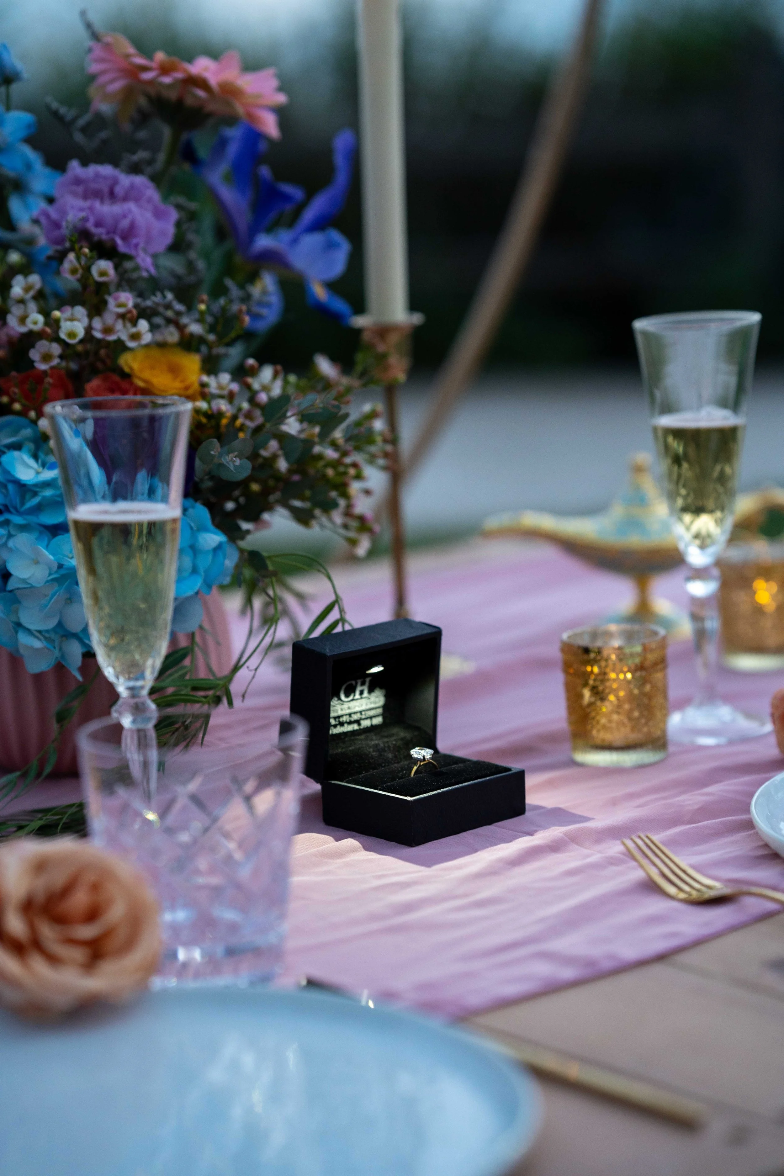 A close-up of a wedding or special event table setting with a pink table runner, gold cutlery, champagne glasses filled with champagne, a silver tray, candle holders, and a bouquet of colorful flowers in the background. A black jewelry box with a rin