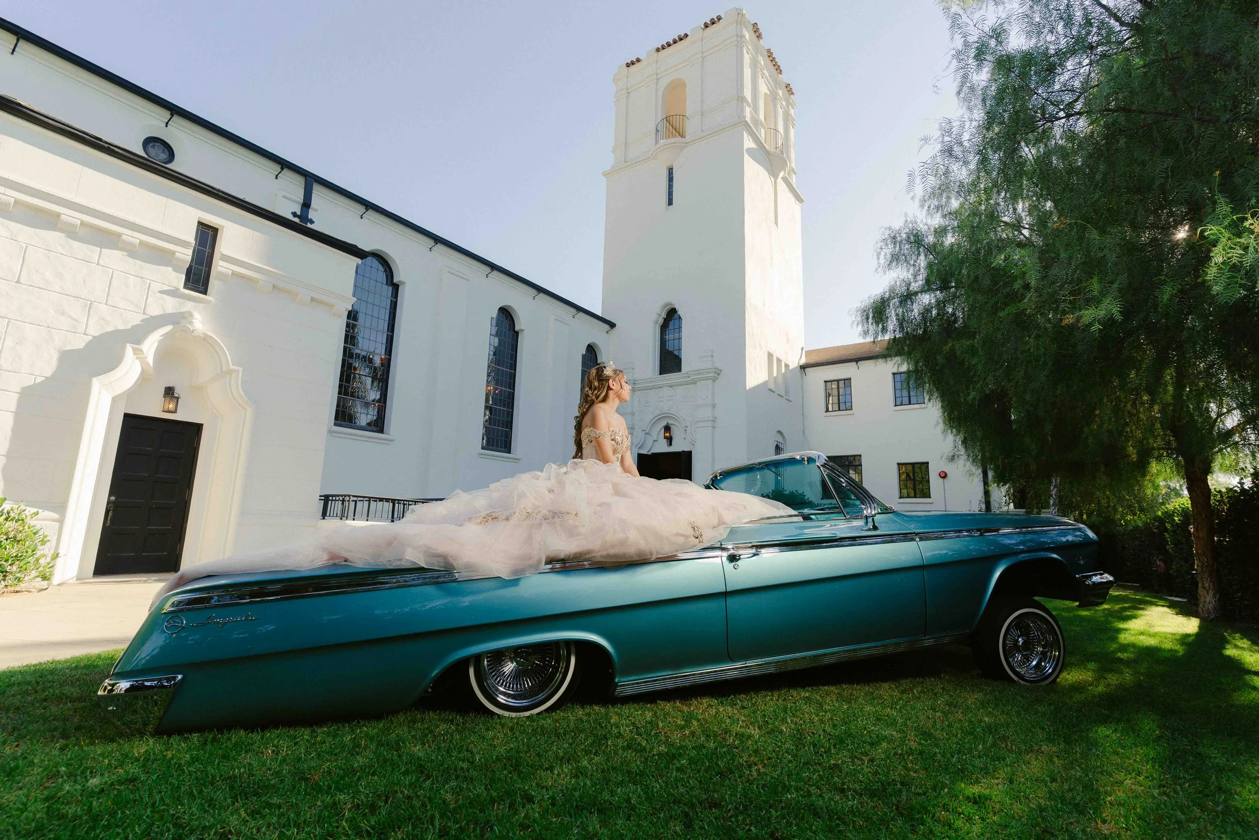 Bride in a wedding gown sitting on a vintage turquoise convertible car parked on a green lawn in front of a white building with tall arched windows and a tower, with trees and clear sky in the background.