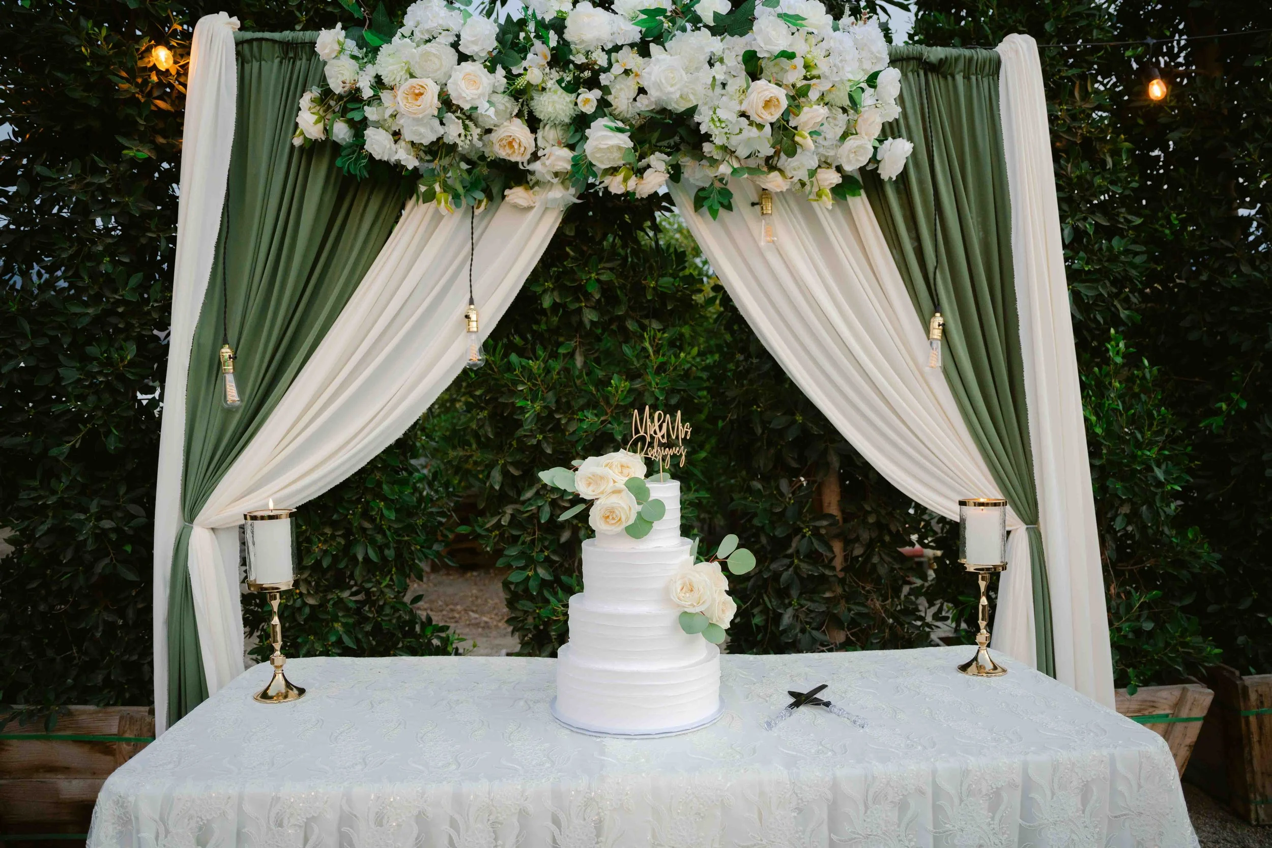 Wedding cake with white roses and eucalyptus on a decorated table under a floral arch with white and green drapes, surrounded by greenery.
