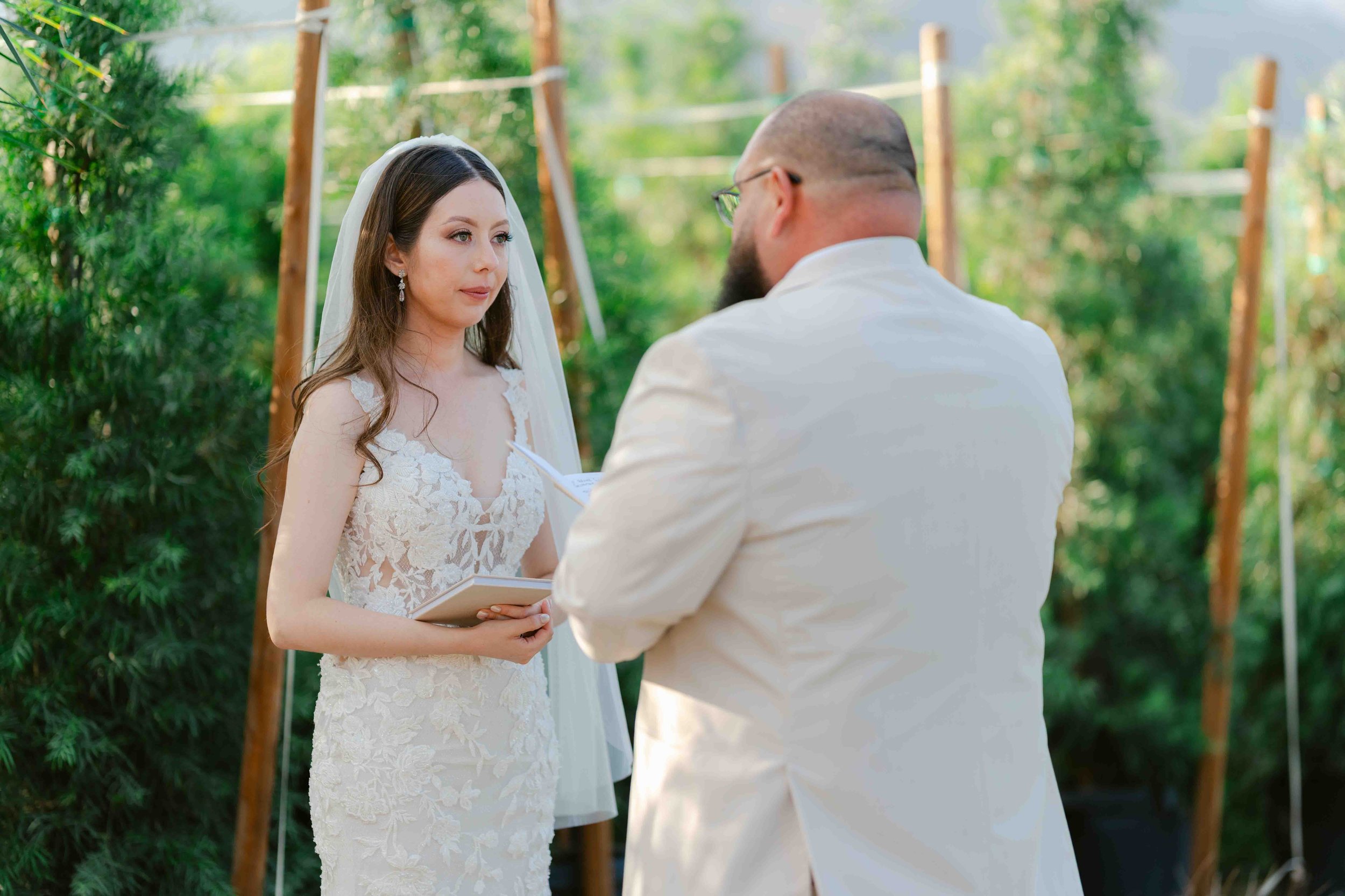 A bride and groom during an outdoor wedding ceremony. The bride wears a white lace wedding dress and veil, holding a small book. The groom wears a white jacket and glasses, facing her. Green trees and wooden arch are in the background.
