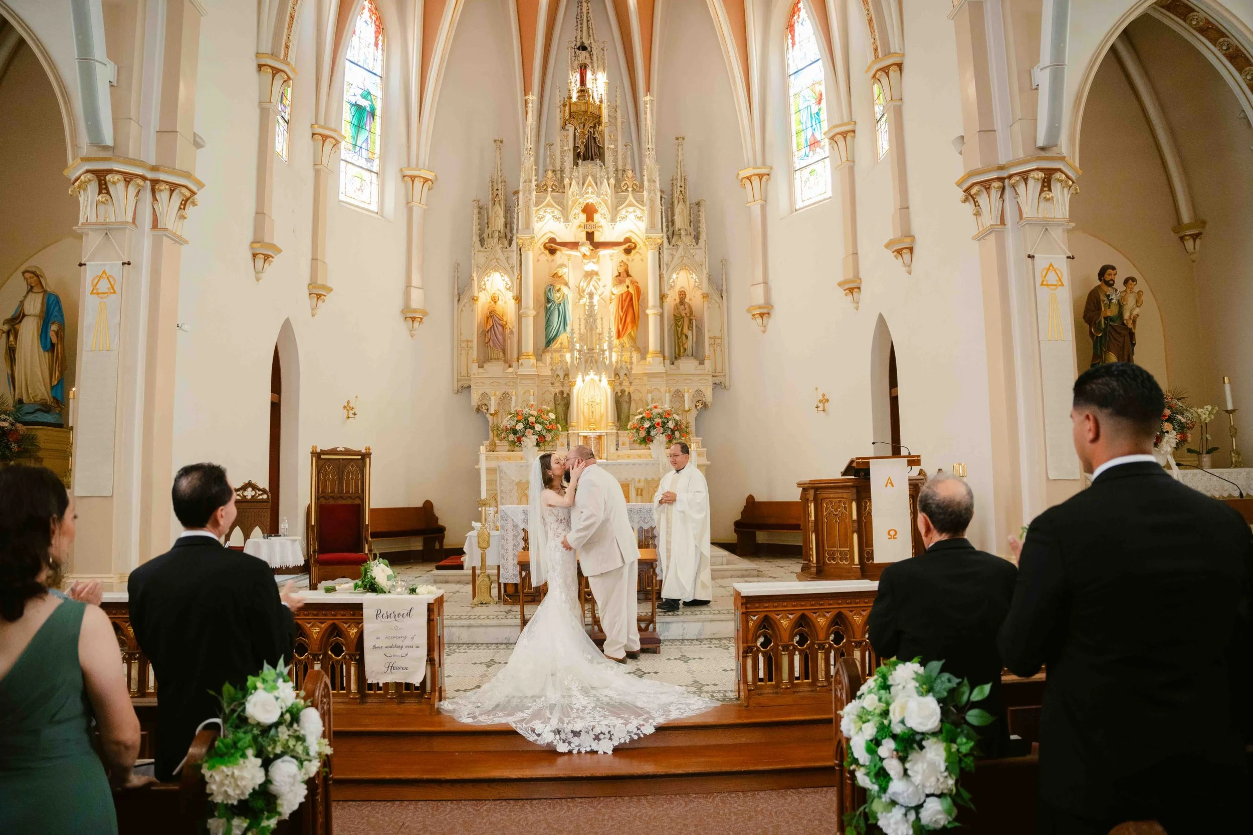A wedding ceremony taking place inside a church with the bride and groom at the altar, surrounded by four guests, a priest, and religious statues and stained glass windows.