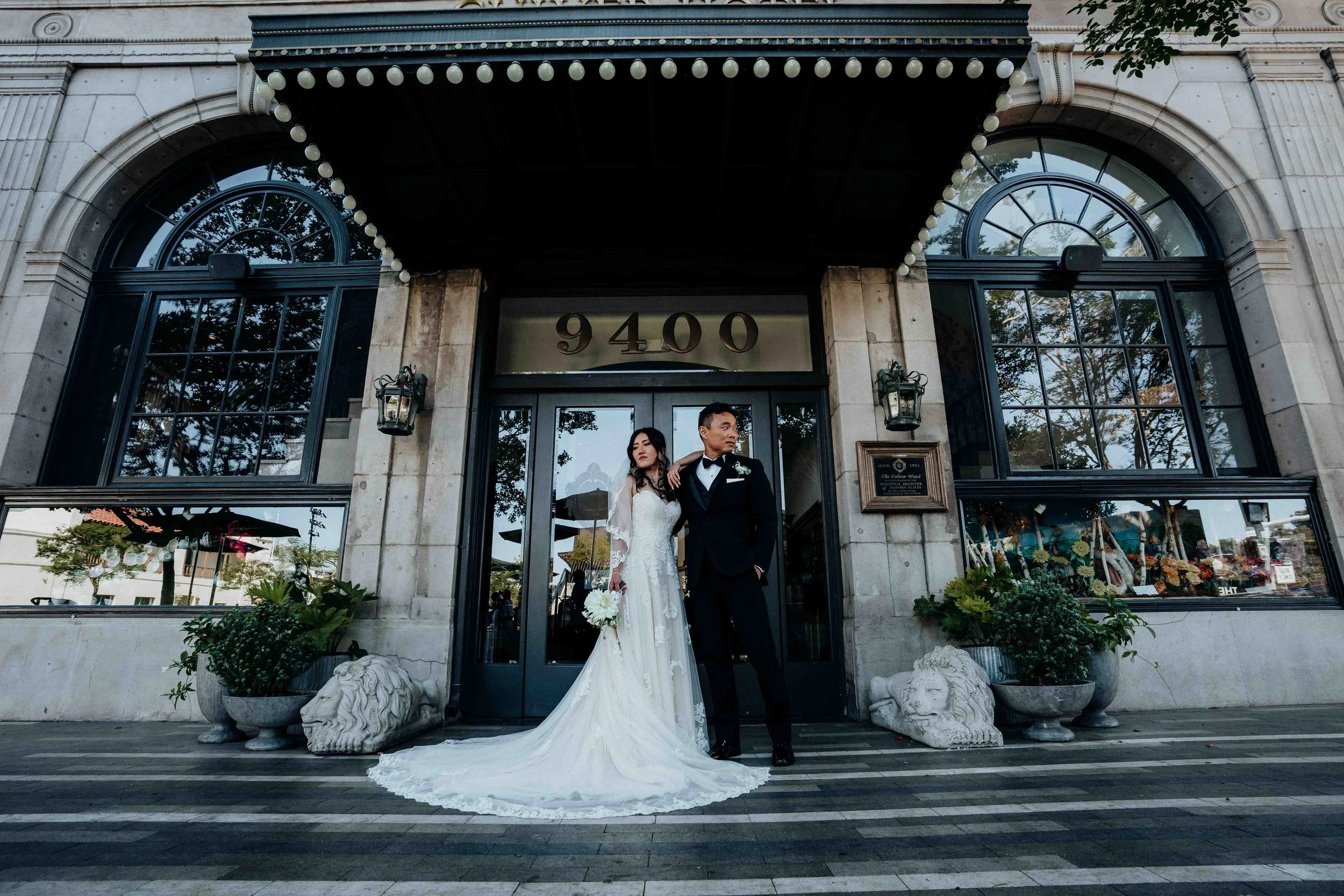 A bride and groom in wedding attire standing in front of a building with the number 9400 above the entrance, surrounded by potted plants and lion statues.