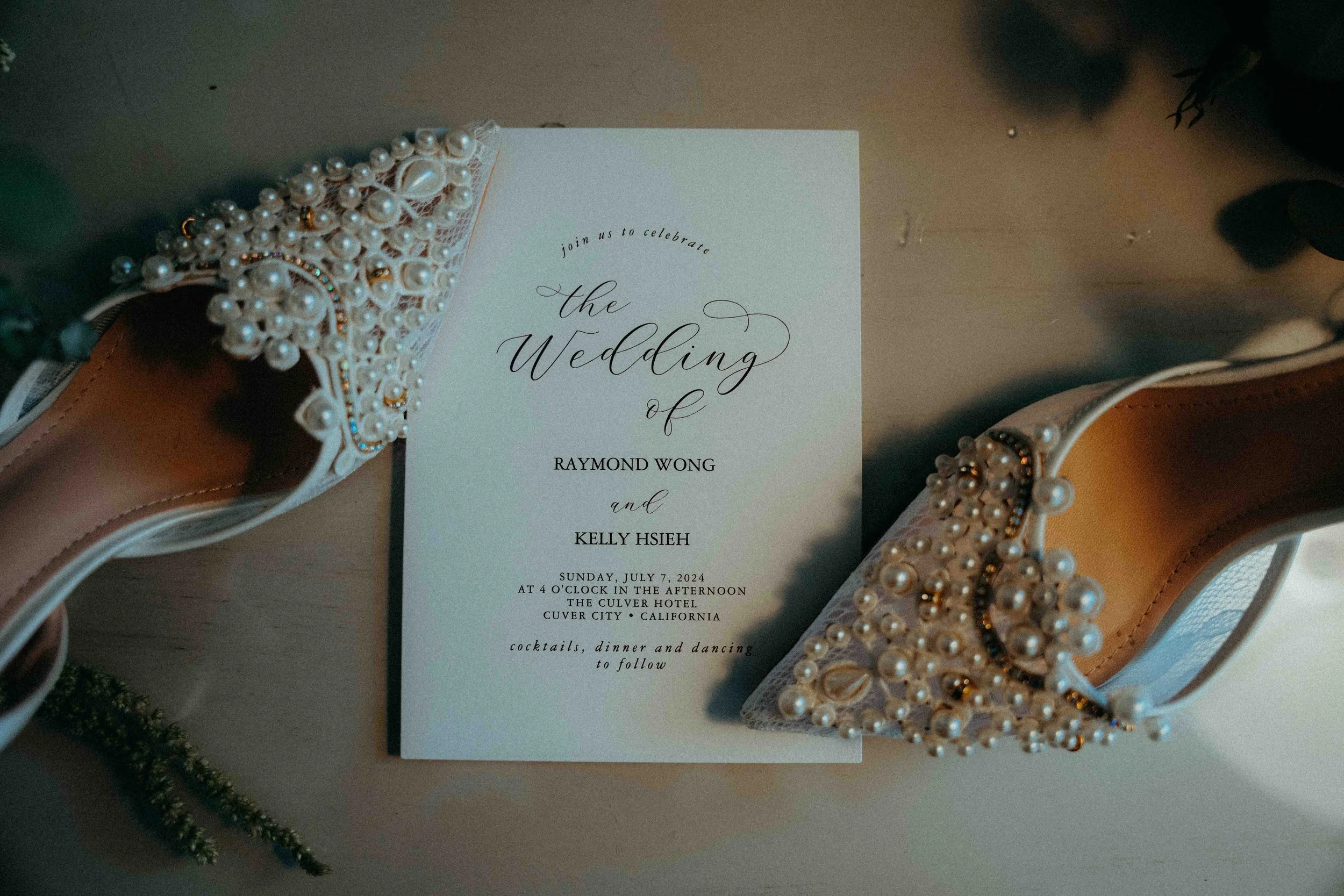 Wedding invitation with pearl-embellished high heels on either side. The invitation is for the wedding of Raymond Wong and Kelly Hsieh, scheduled for July 7, 2024, at the Culver Hotel in Culver City, California.