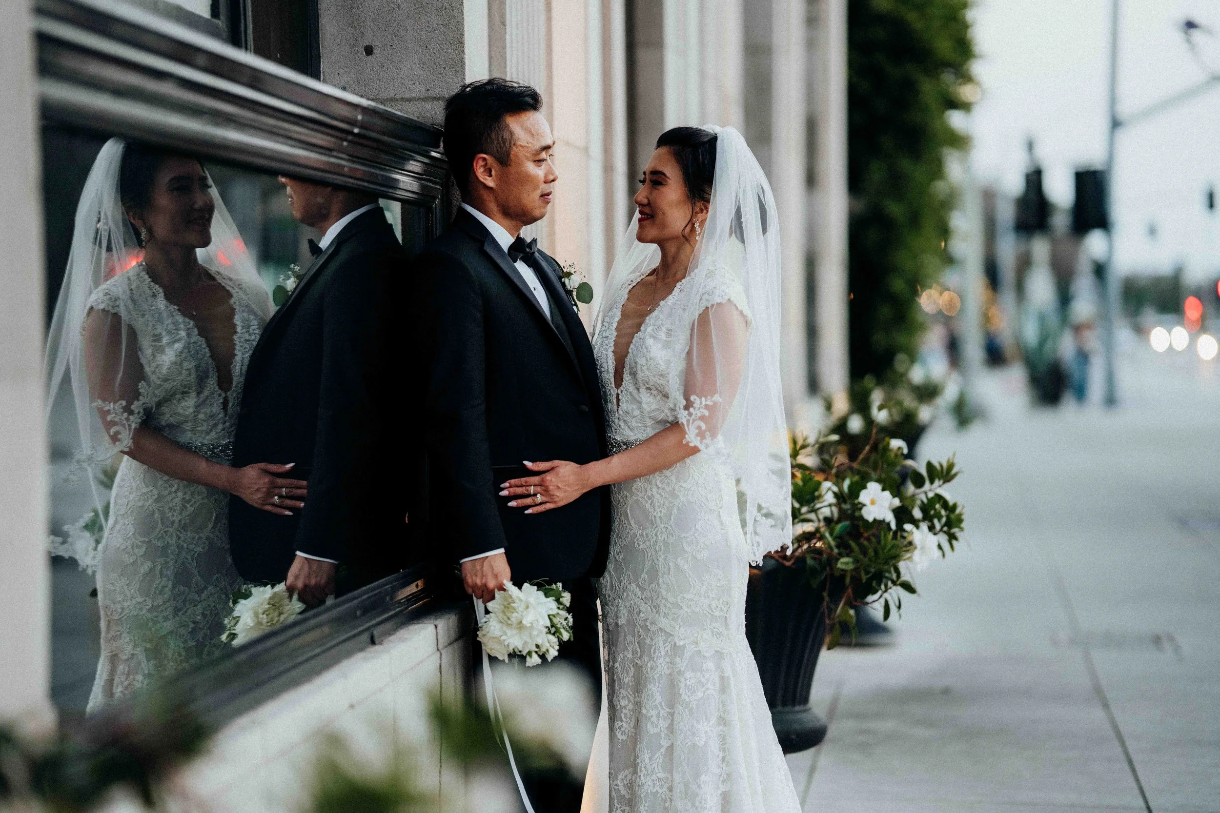 A bride and groom in wedding attire standing close together outside on a city sidewalk, with the bride touching the groom's chest and both smiling.