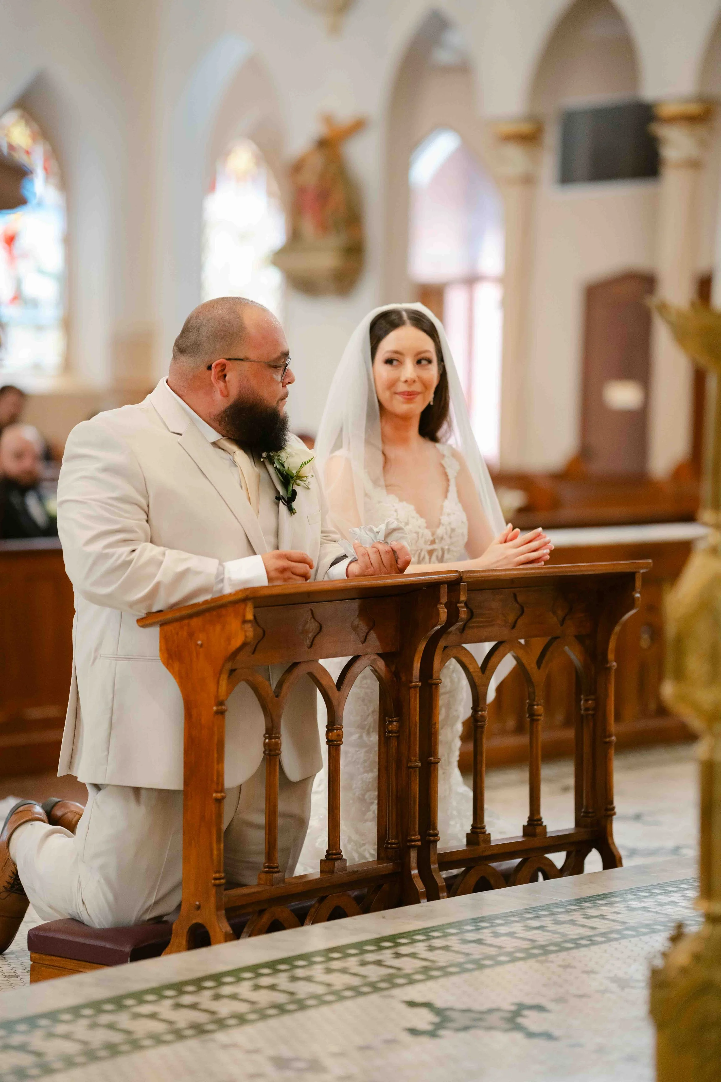 Kneeling bride and groom praying at their wedding ceremony in a church, with wooden pews and religious statues in the background.