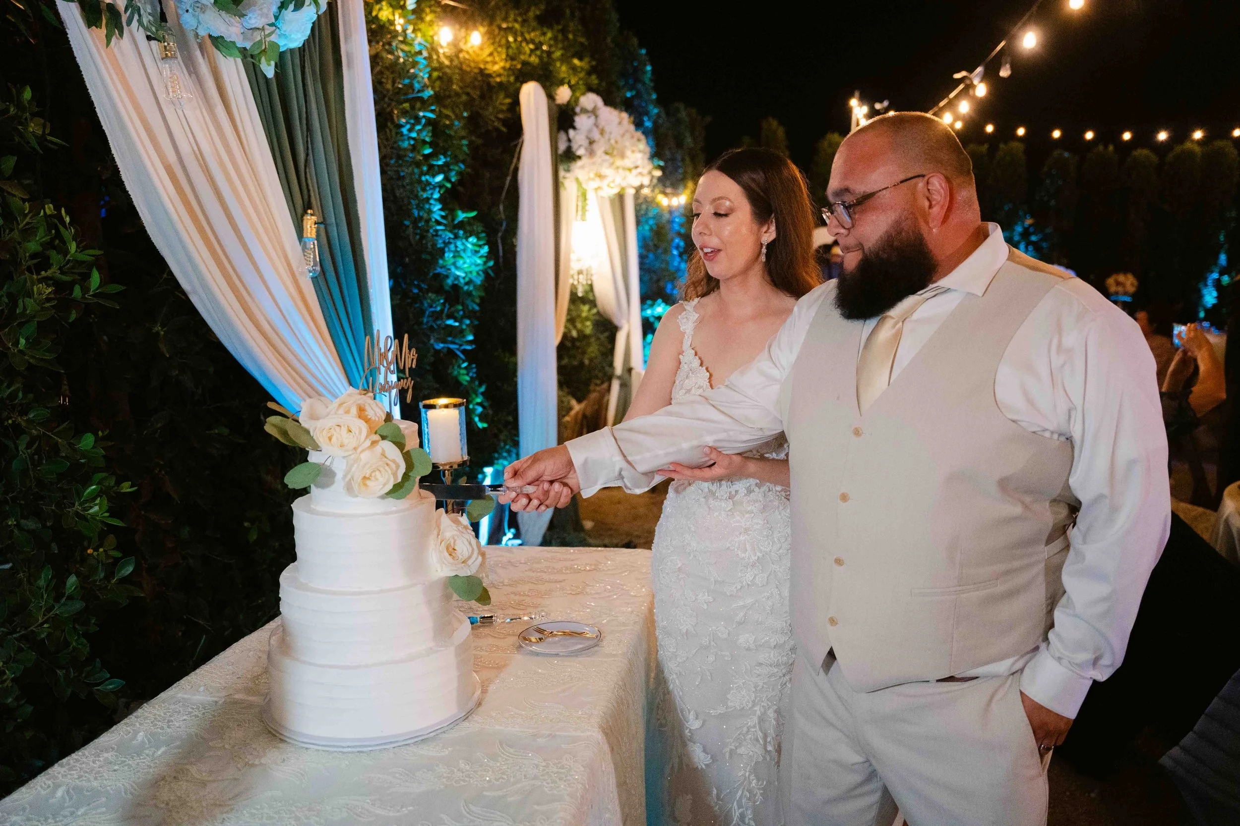 A bride and groom in wedding attire cutting a wedding cake together outdoors at night, decorated with white roses and greenery, under string lights and draped fabric.