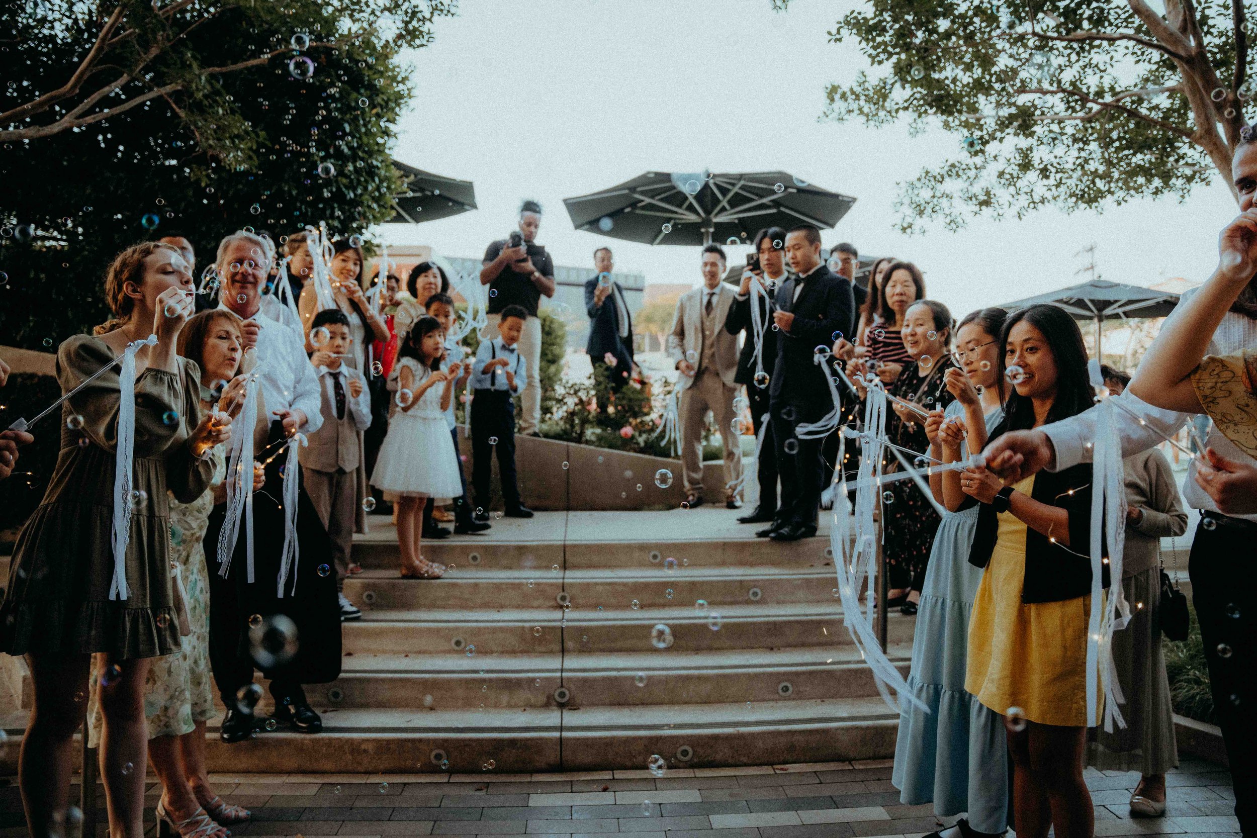 Guests at a wedding celebration playing with bubbles on outdoor steps with umbrellas and trees in the background.