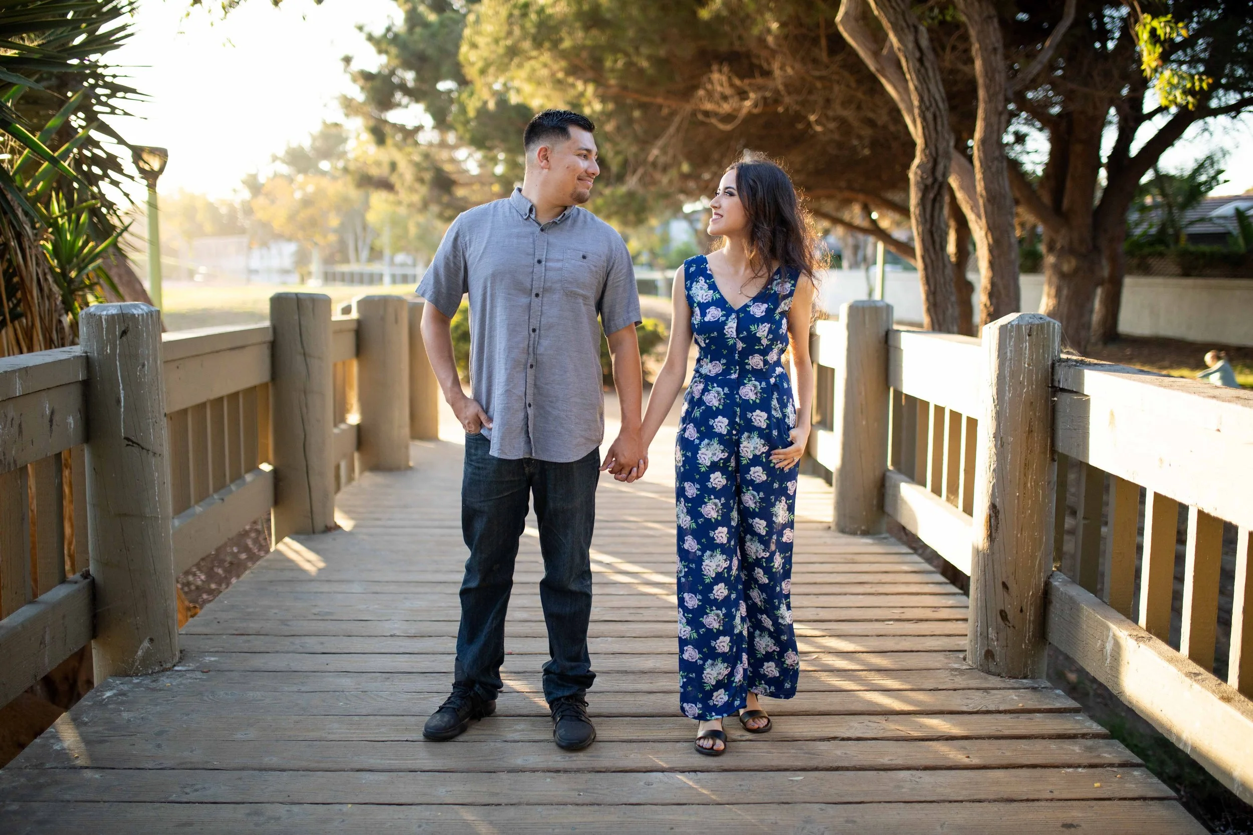 A young couple holding hands and walking on a wooden bridge in a park, smiling at each other, with trees and sunlight in the background.