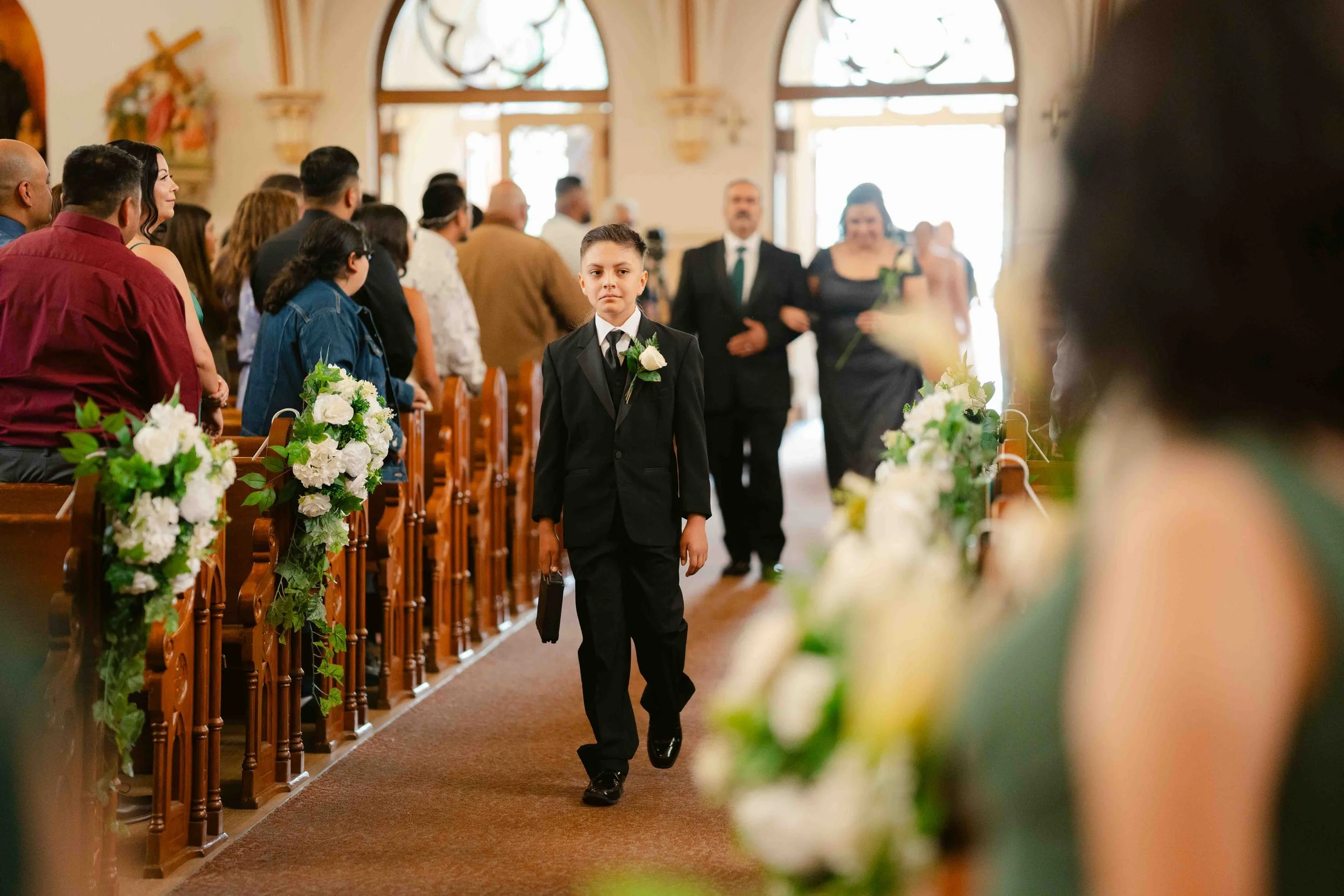 A young boy in a black tuxedo walking down the aisle of a church decorated with white flowers and green foliage, with guests seated on both sides, during a wedding ceremony.