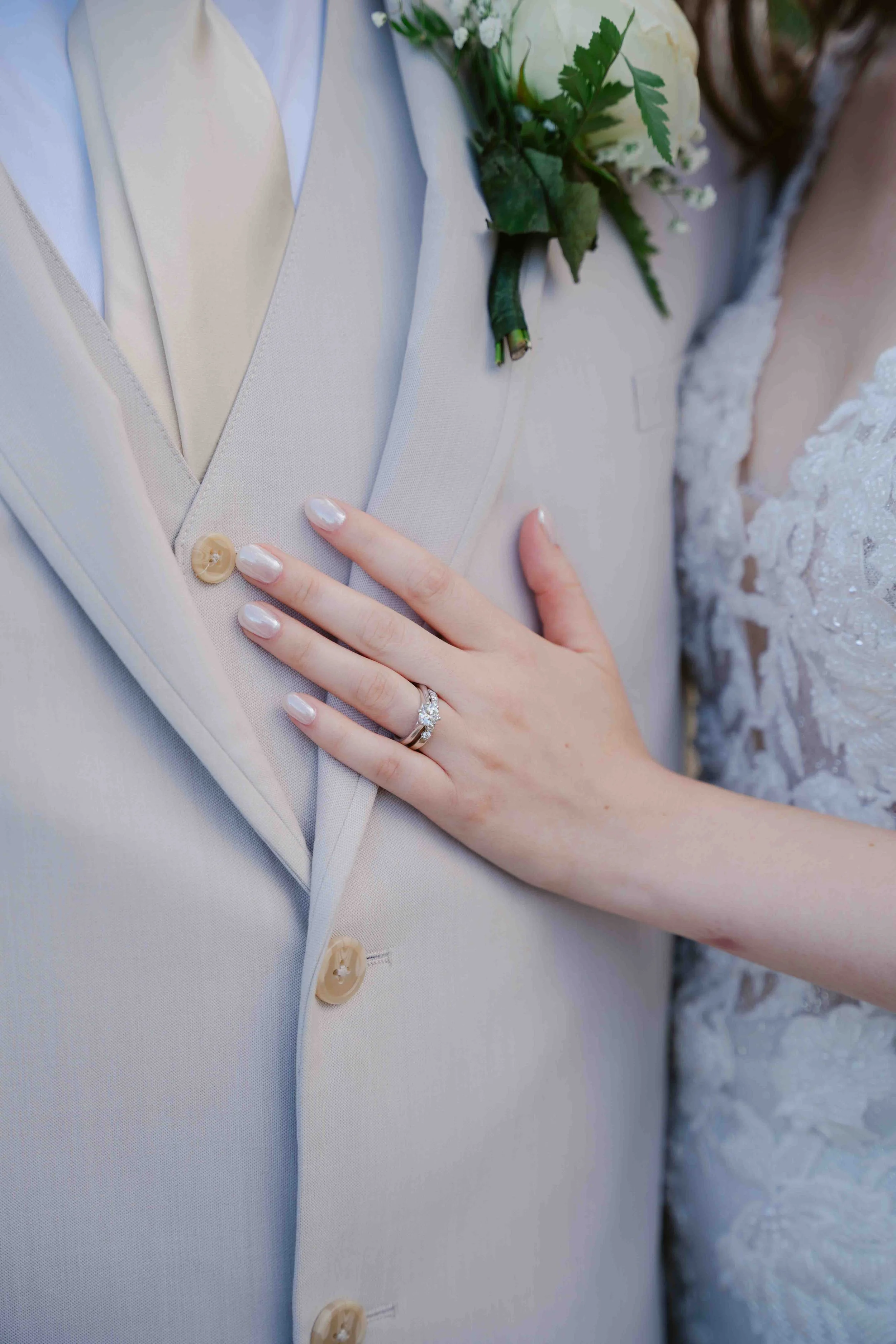 Close-up of a bride's hand resting on a groom's chest, showing her wedding ring and engagement ring. The groom is wearing a light-colored suit with a boutonnière of white flowers and greenery. The bride's dress has lace details.