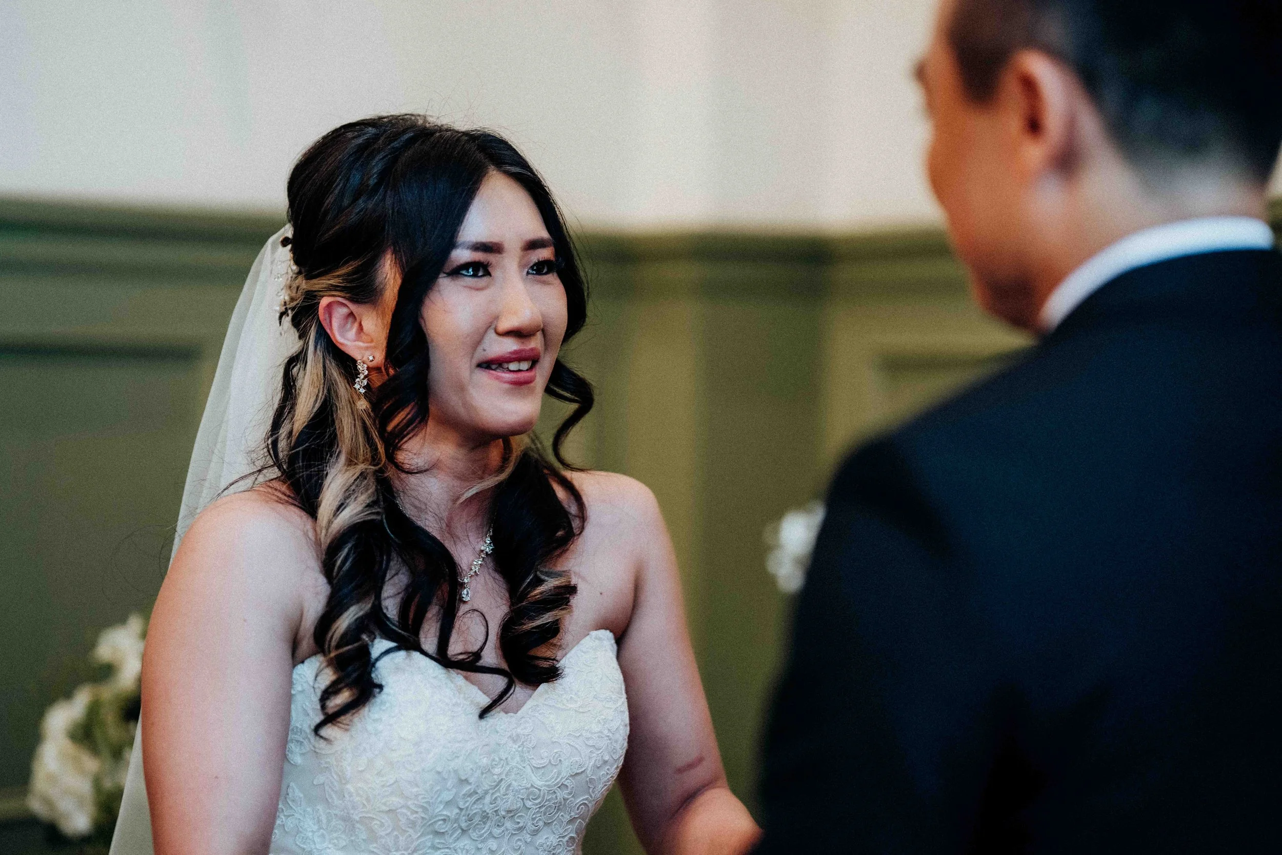 A bride with long dark hair and a white wedding dress looks emotionally at a groom during their wedding ceremony.
