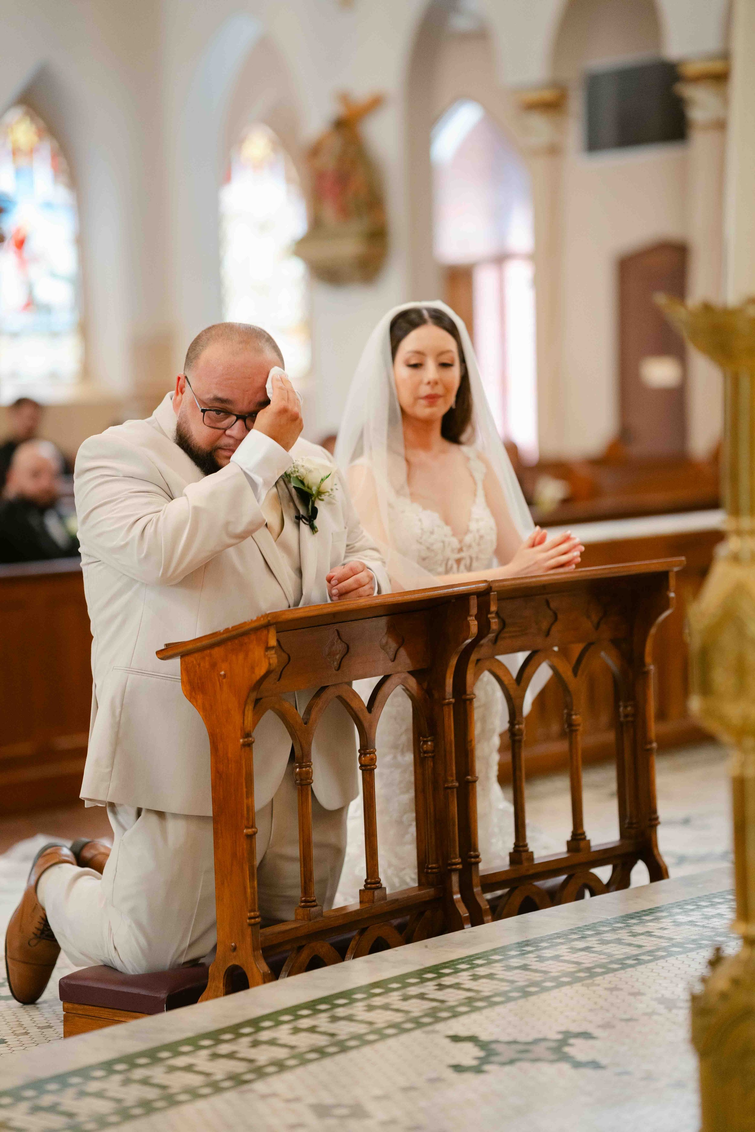 A man and woman kneeling in prayer during a wedding ceremony inside a church, with stained glass windows in the background.