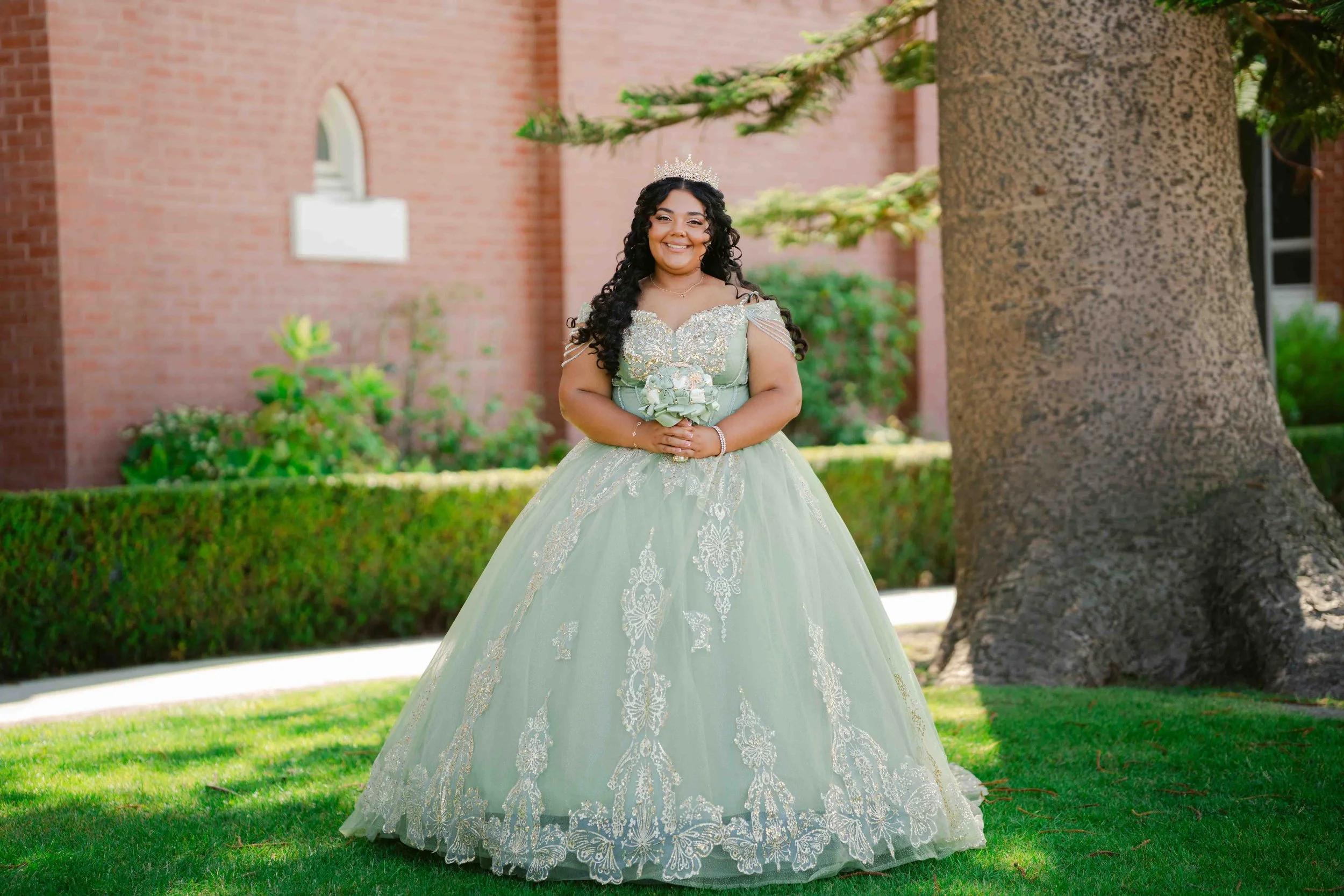 Young woman dressed in an elegant, light green quincenera dress with intricate lace details, wearing a tiara, holding a small bouquet, standing outdoors next to a large tree with a brick building in the background.