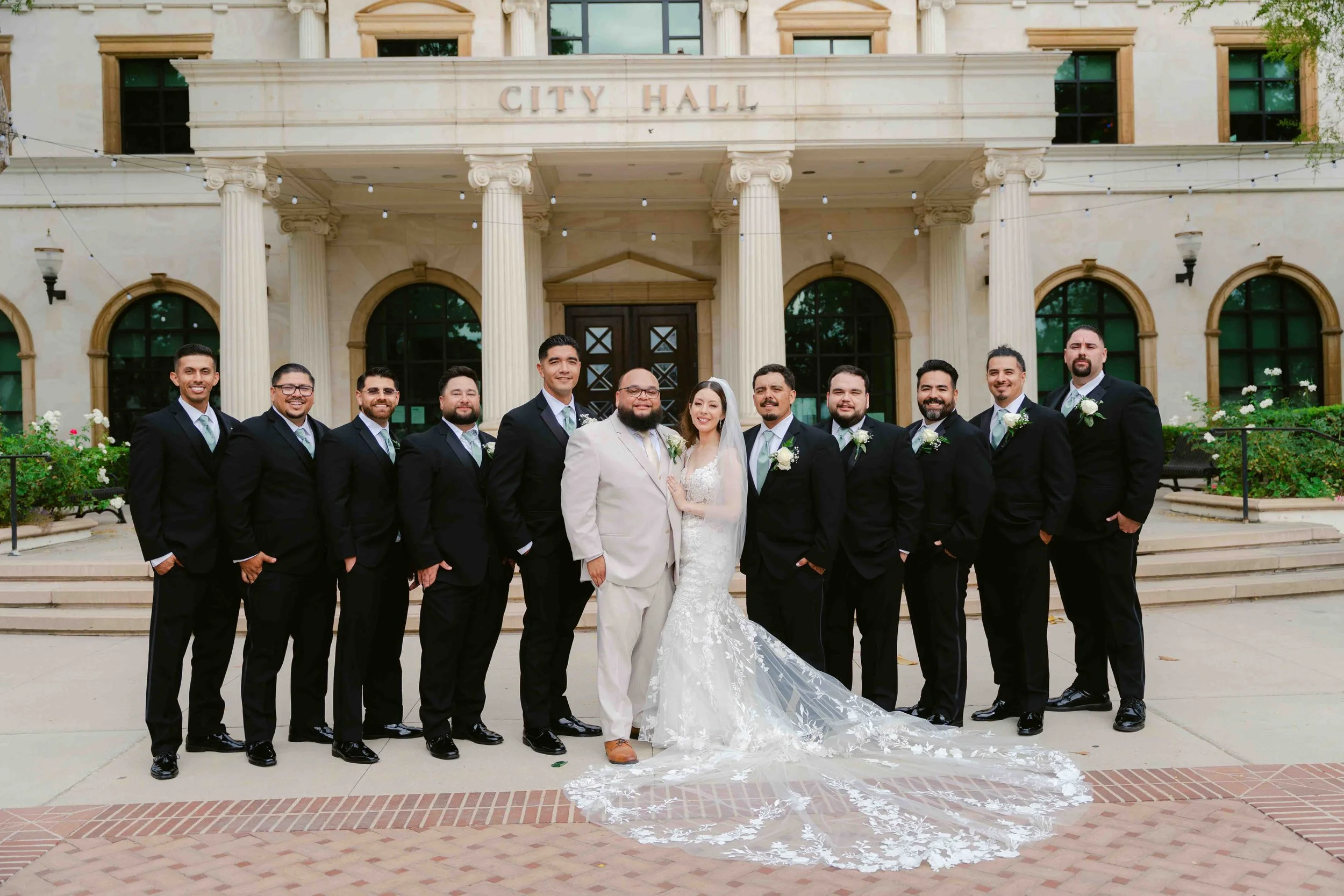 Group of people dressed in wedding attire standing outside a building labeled City Hall, with a bride in a white gown and veil, and groom in a light-colored suit, surrounded by groomsmen in black suits.
