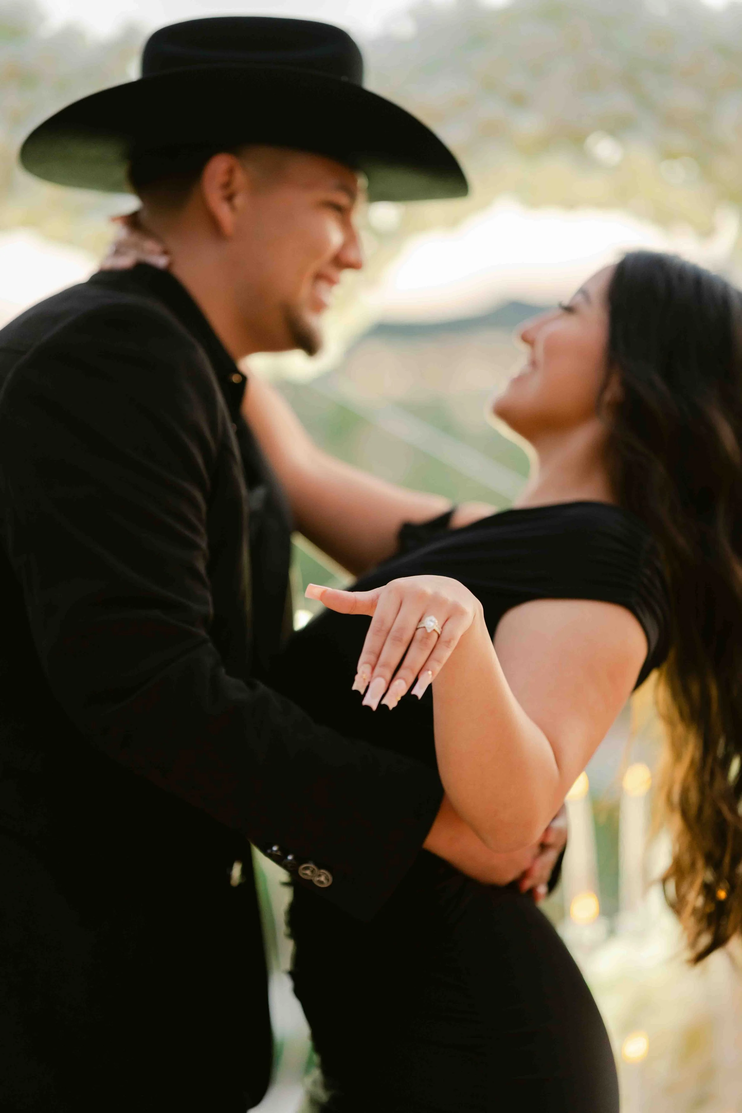 A man and a pregnant woman are dancing together, smiling and looking at each other. The man is wearing a black hat and a suit, and the woman is wearing a black dress and has long dark hair.