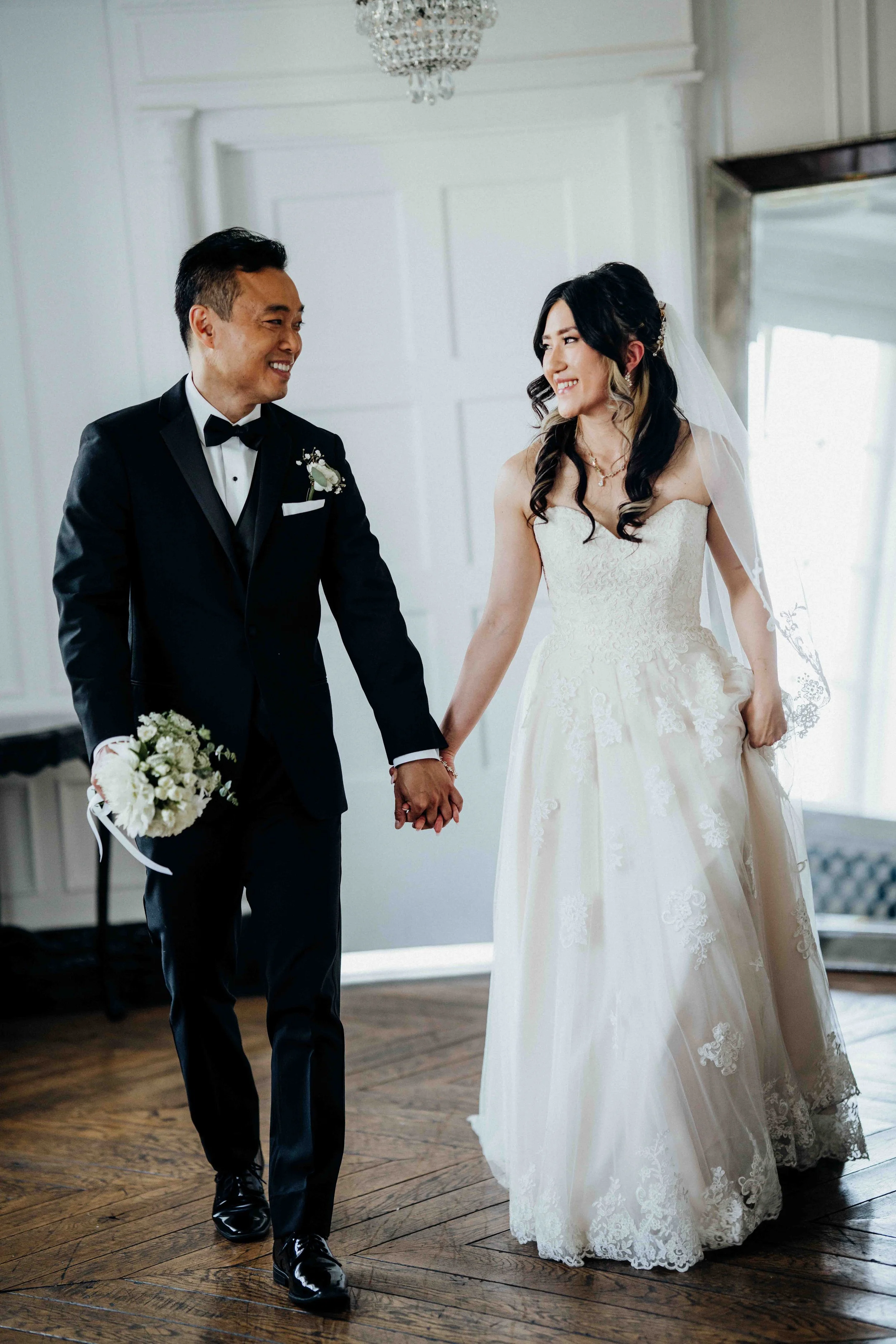 A bride in a white wedding gown and a groom in a black tuxedo holding hands and smiling at each other inside a room with white walls and wooden floors.
