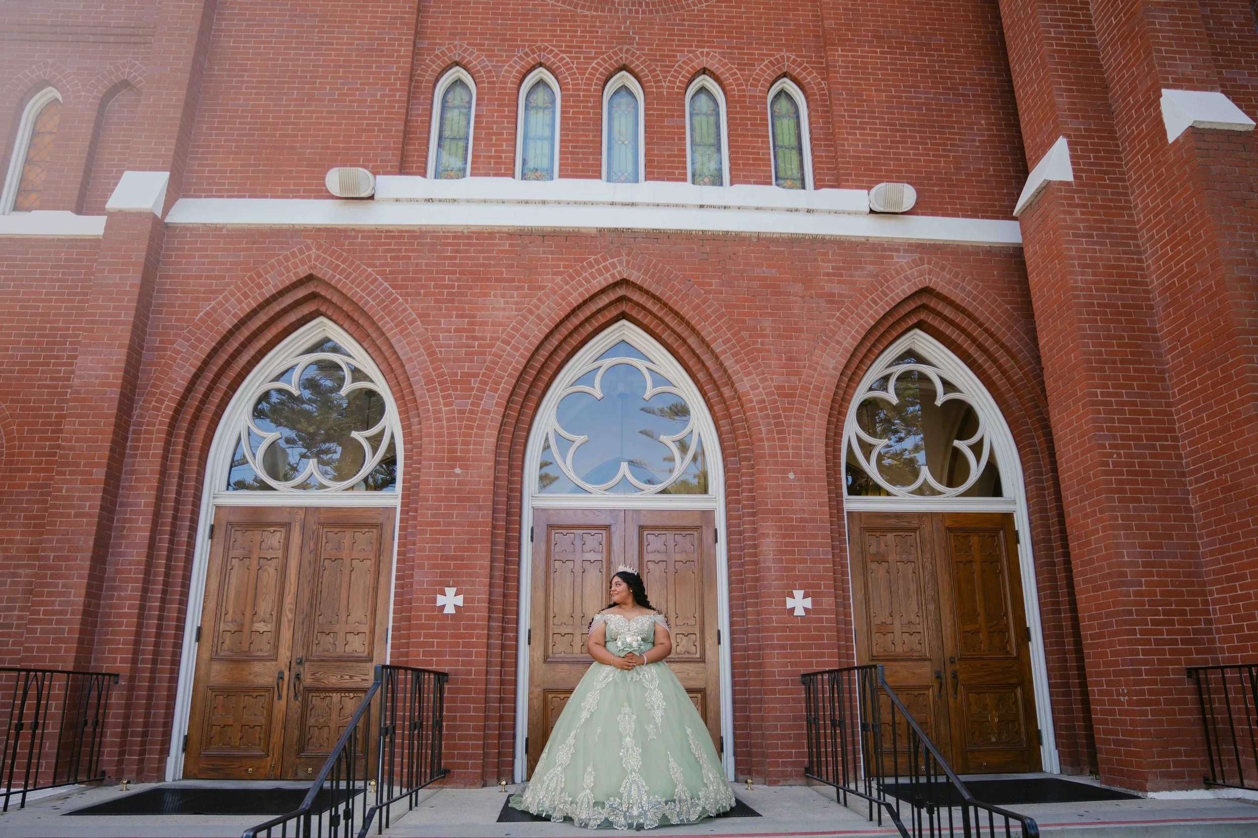 A young woman in a light green, ornate formal dress with lace and embroidery stands in front of a red brick church with large wooden doors and decorative stained glass windows. She is wearing a tiara and looking to the side.