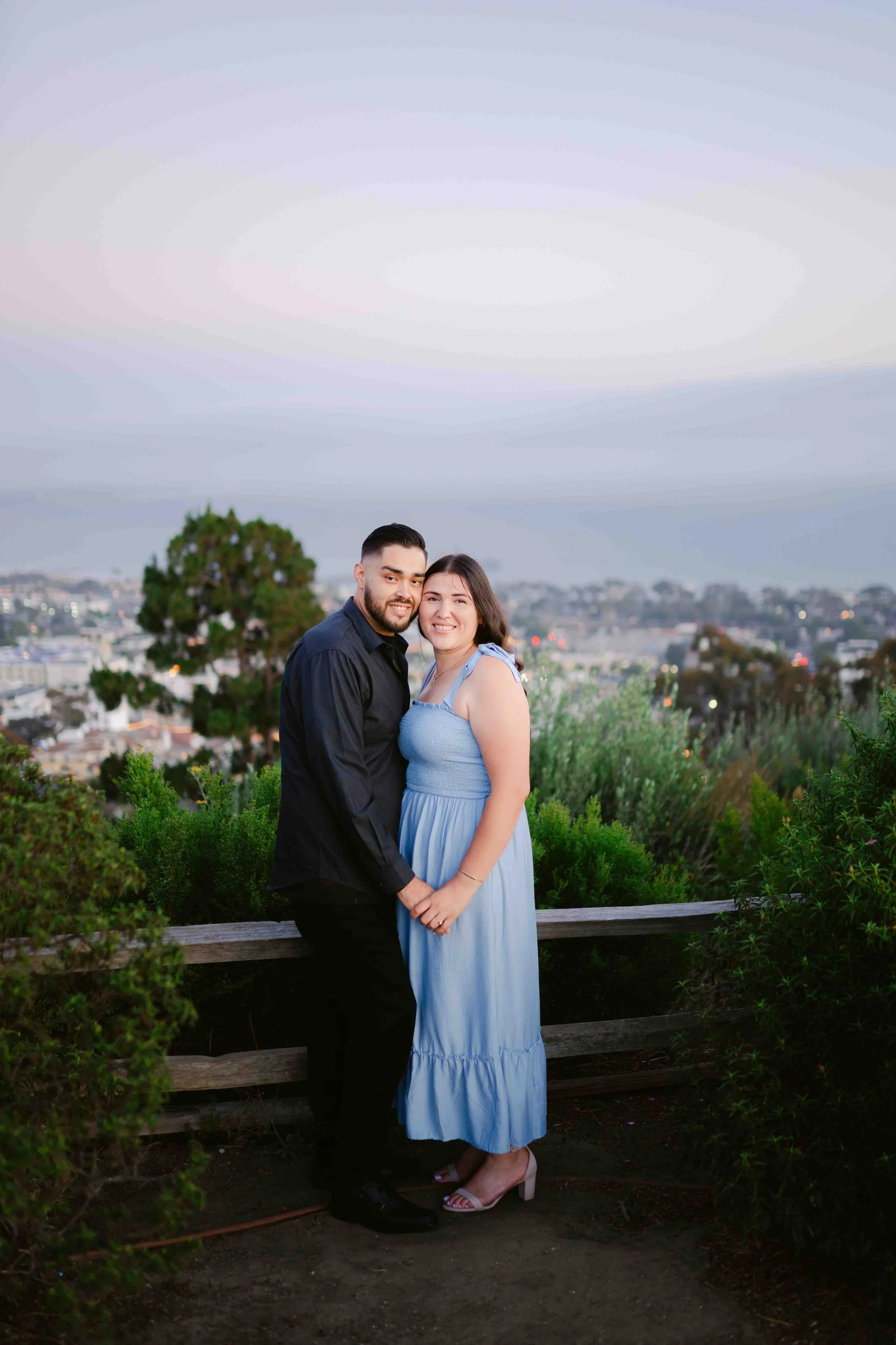A young couple standing close together outdoors on a city overlook during dusk, smiling for a photo. The man has dark hair and a beard, wearing a dark shirt and black pants. The woman has shoulder-length dark hair, wearing a light blue dress and ligh