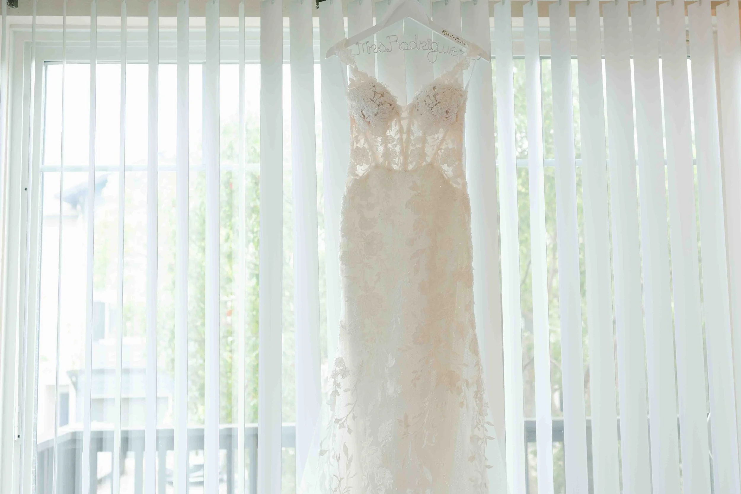 Wedding dress hanging on a hanger in front of window with vertical blinds.