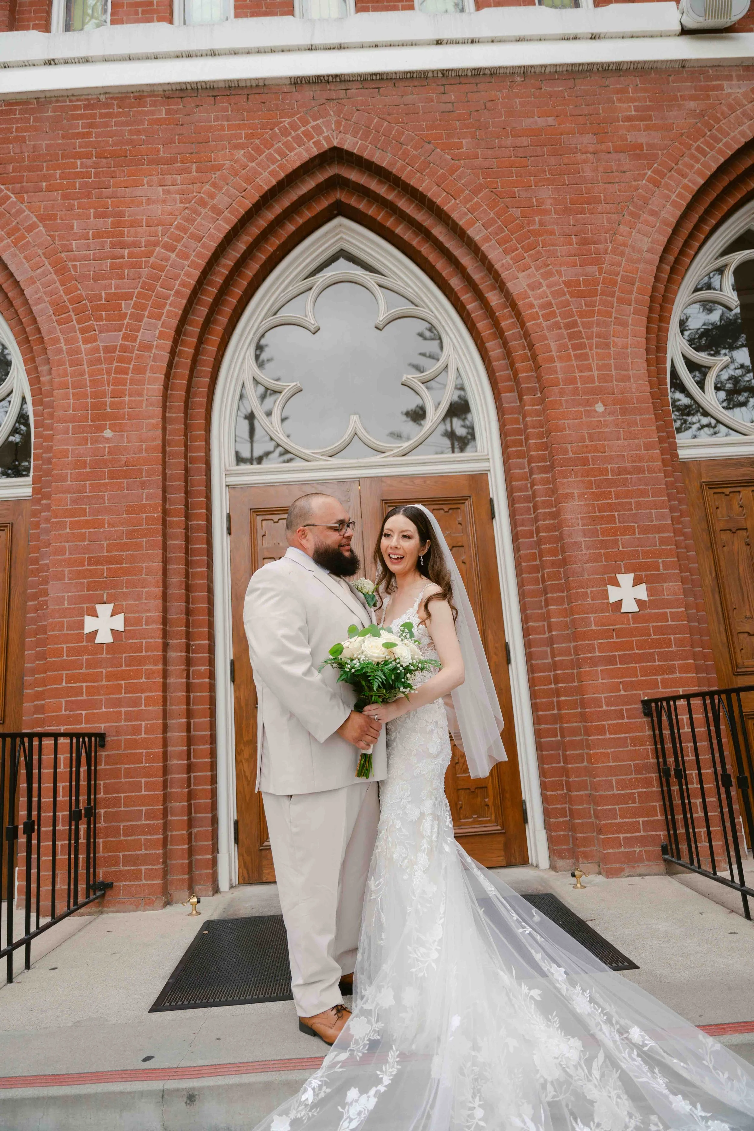 A bride and groom standing together outside a red brick church with wooden double doors, smiling and holding a bouquet of white flowers.