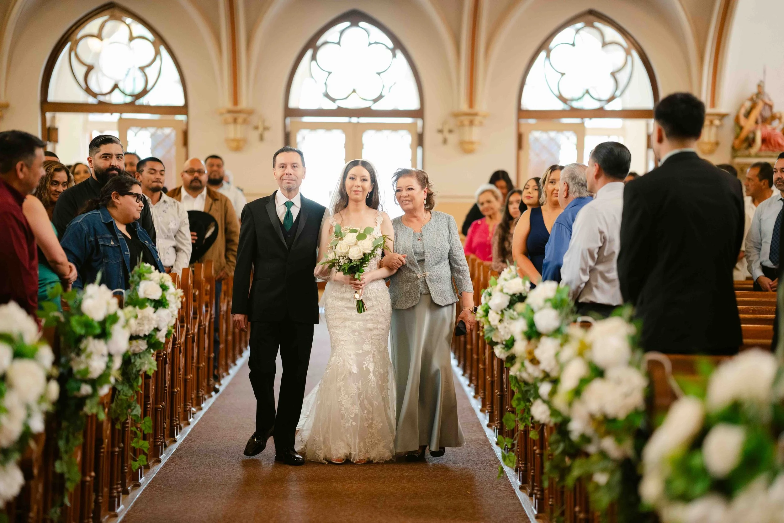 A bride walking down the aisle with her mother, surrounded by wedding guests seated in a church decorated with white flowers.