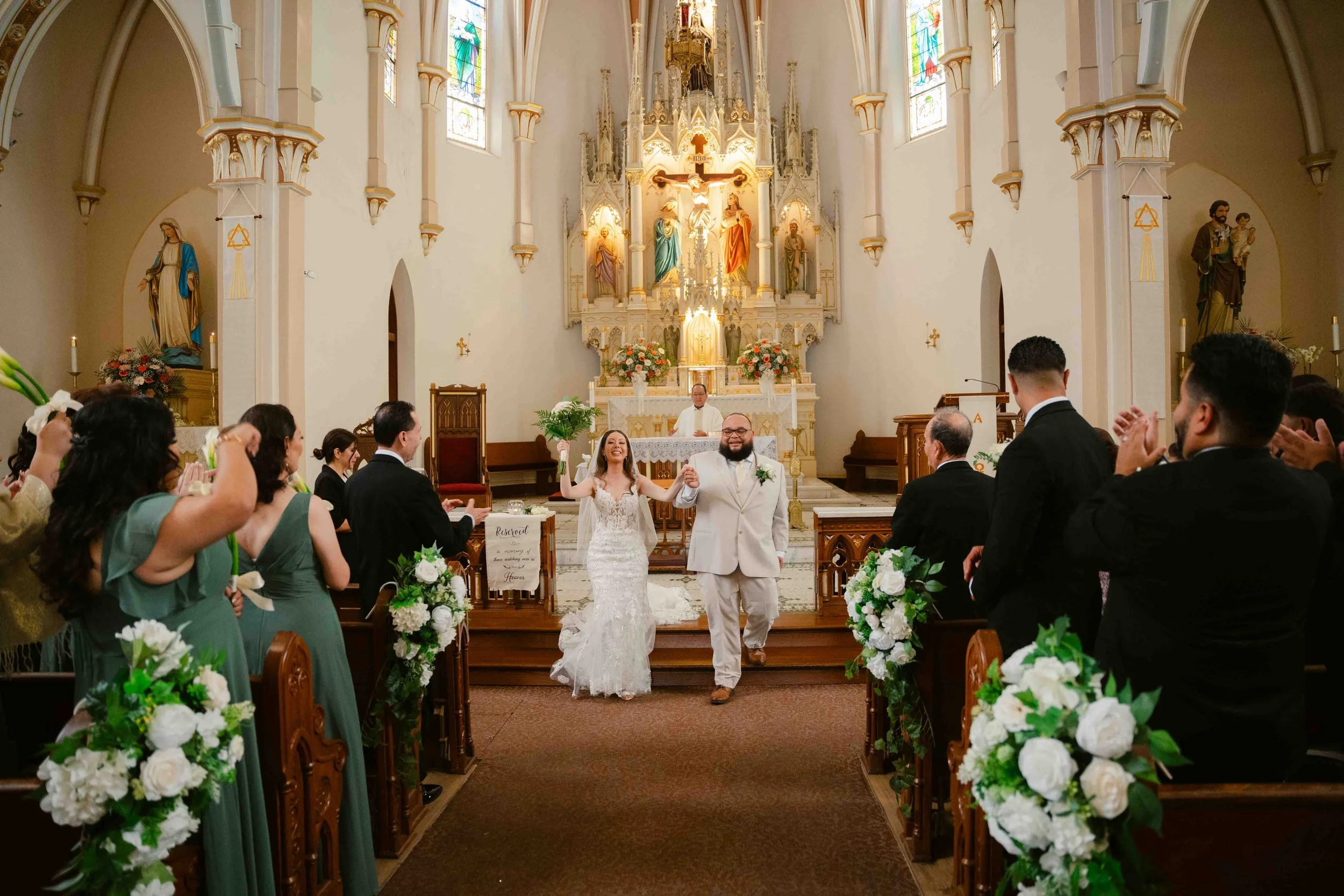 A newlywed couple walking down the aisle in a church, holding hands, with the bride holding a bouquet, while guests on both sides clapping and celebrating, and a priest behind the altar.