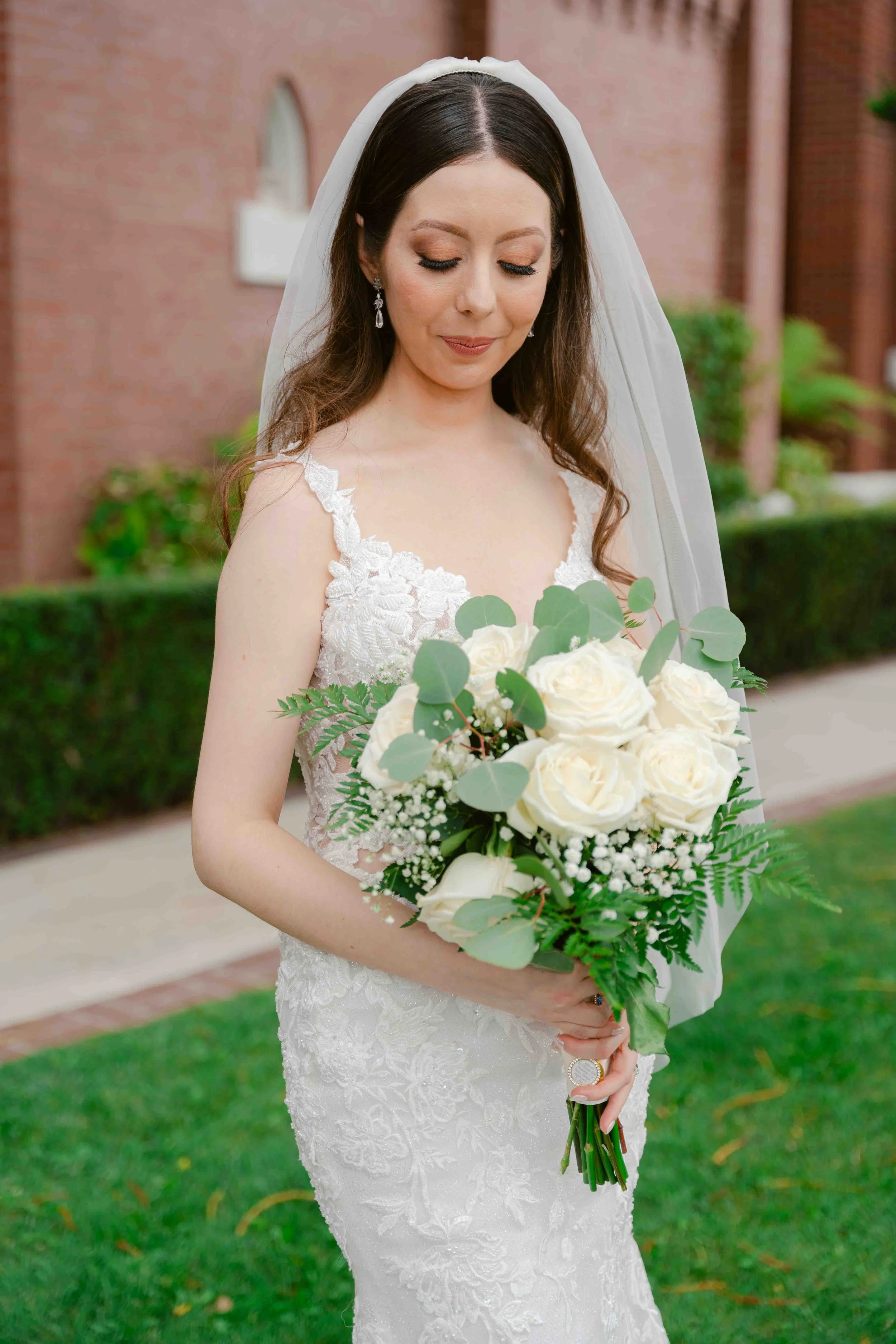 A bride in a white lace wedding gown holding a bouquet of white roses and greenery, standing outdoors with a brick building and shrubs in the background.