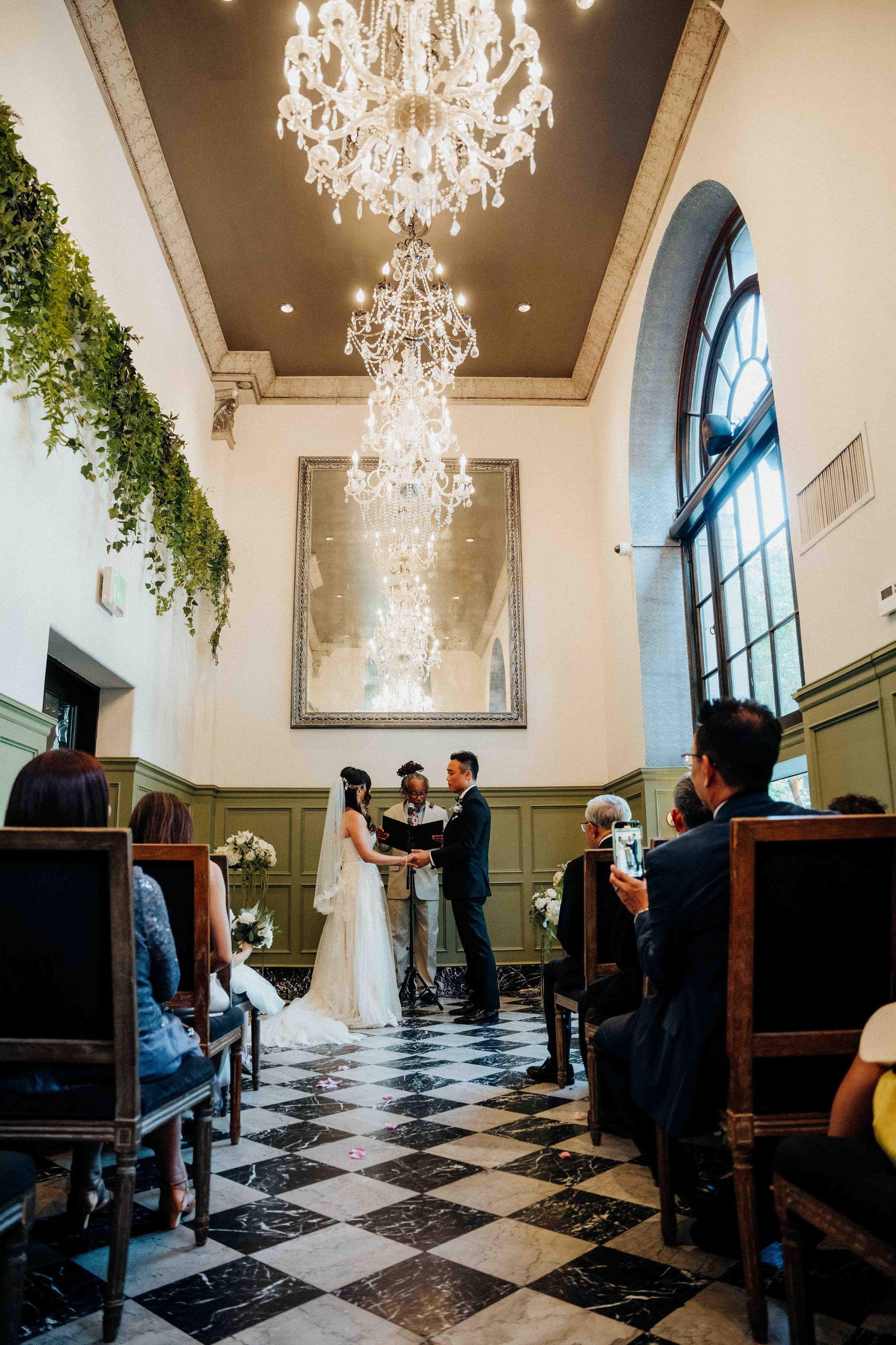 A wedding ceremony with a bride and groom exchanging vows in an elegant indoor venue with chandeliers and a large mirror on the wall.