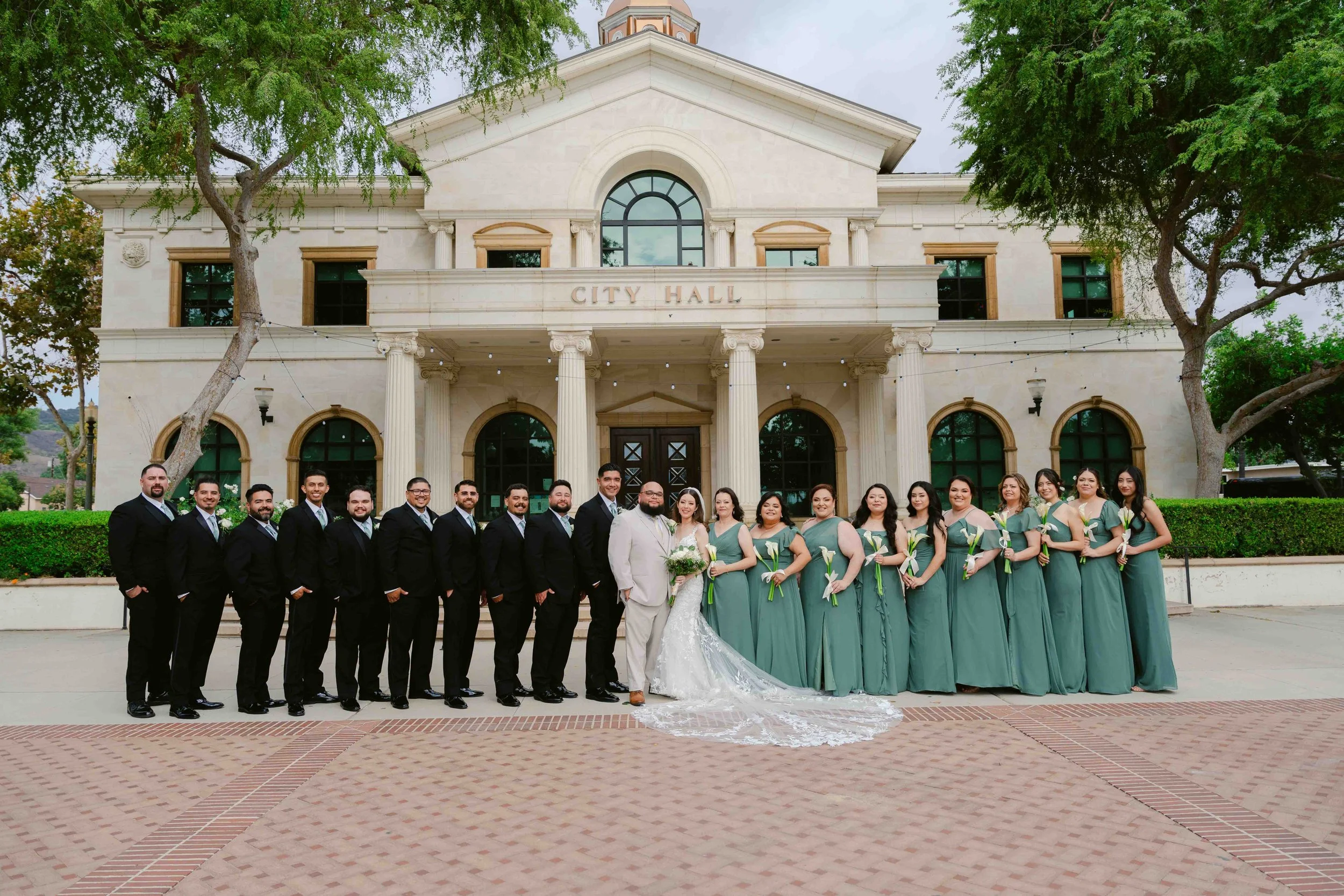 A wedding party standing in front of a city hall building, with men in black suits and women in matching green dresses, some holding white calla lilies, and the bride in a white lace gown with a long train and bouquet, and the groom in a light-colore