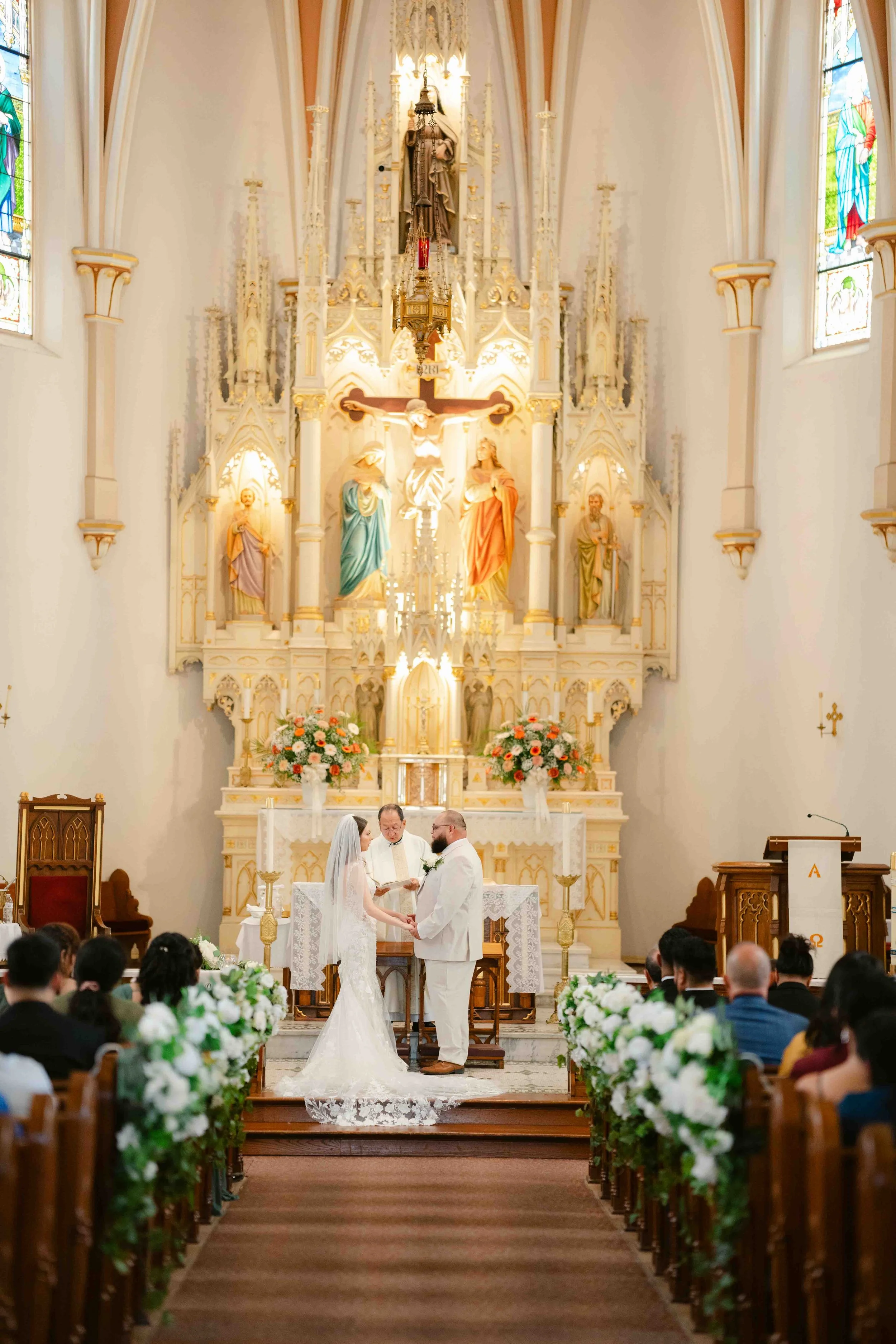 A wedding ceremony taking place inside a church with a bride and groom standing at the altar while a priest officiates. Guests are seated on both sides, and the church features ornate decorations, stained glass windows, and floral arrangements.
