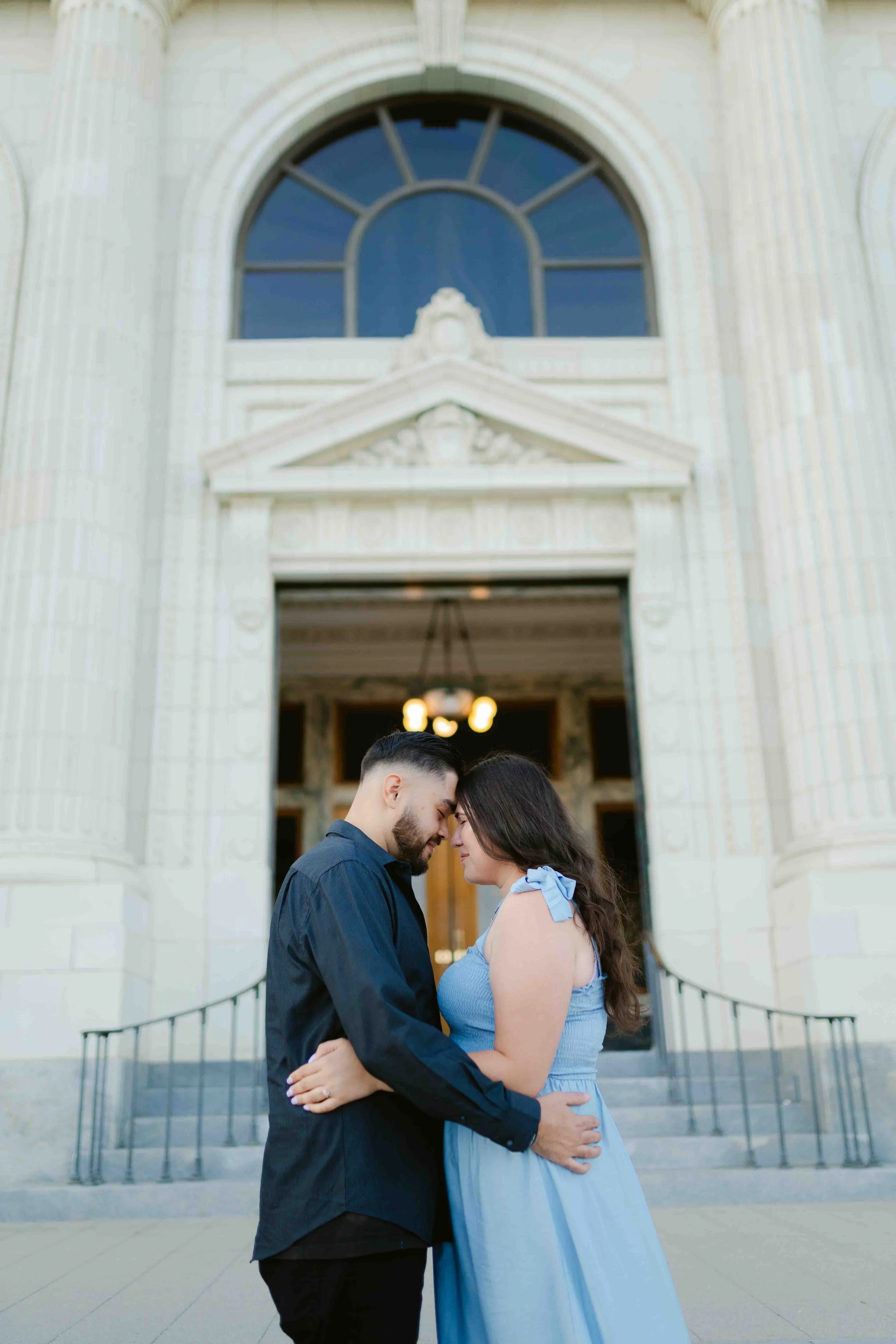 A young couple stands close together, touching foreheads and smiling softly outside a historic building with large arched windows and ornate architectural details. The man wears a dark shirt, and the woman wears a light blue dress with shoulder bows.