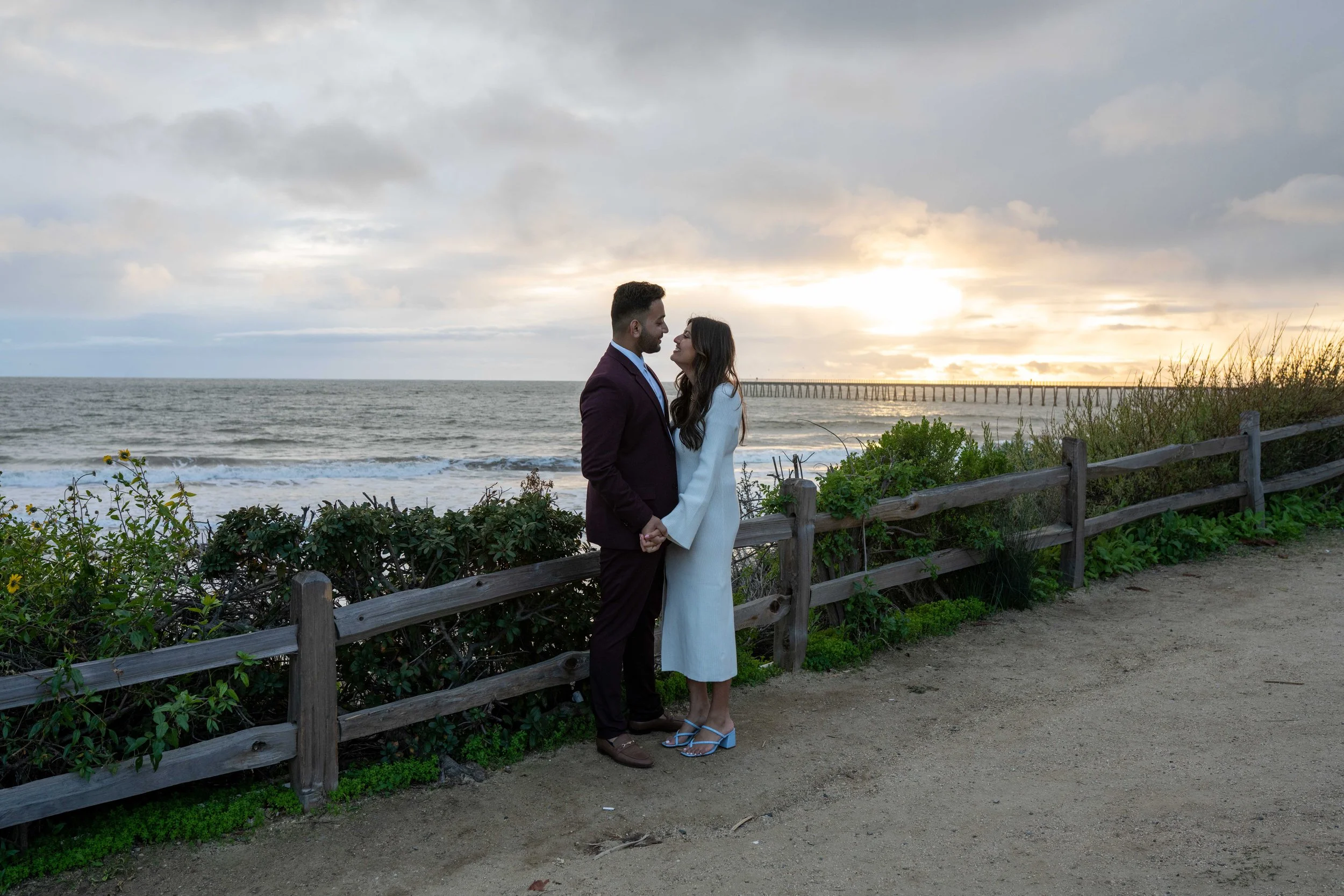 A couple holding hands and looking at each other on a beachside walkway during sunset, with the ocean, a pier, and cloudy sky in the background.