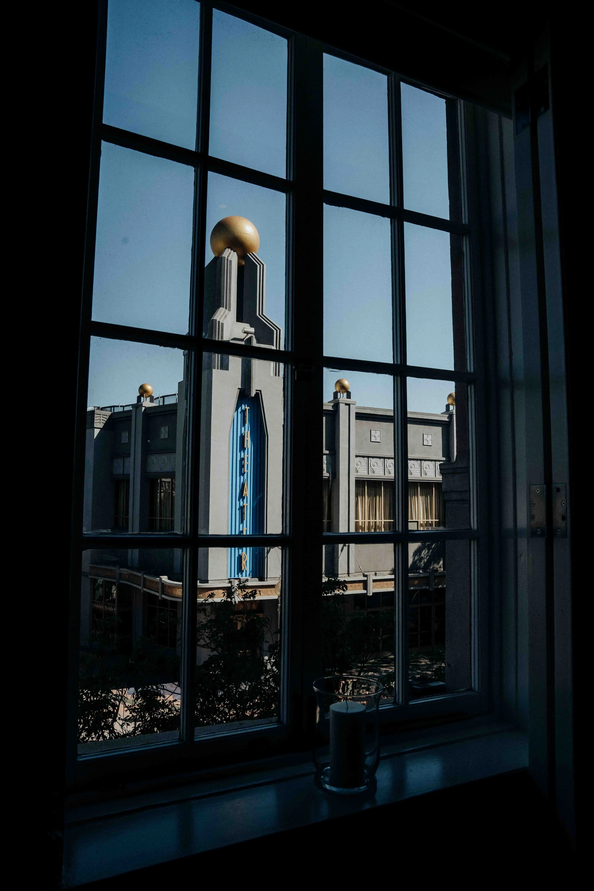 View through a window showing a building with an art deco style, featuring gray walls, gold decorative spheres on top, and a blue vertical sign with gold letters that reads 'THEATER'.
