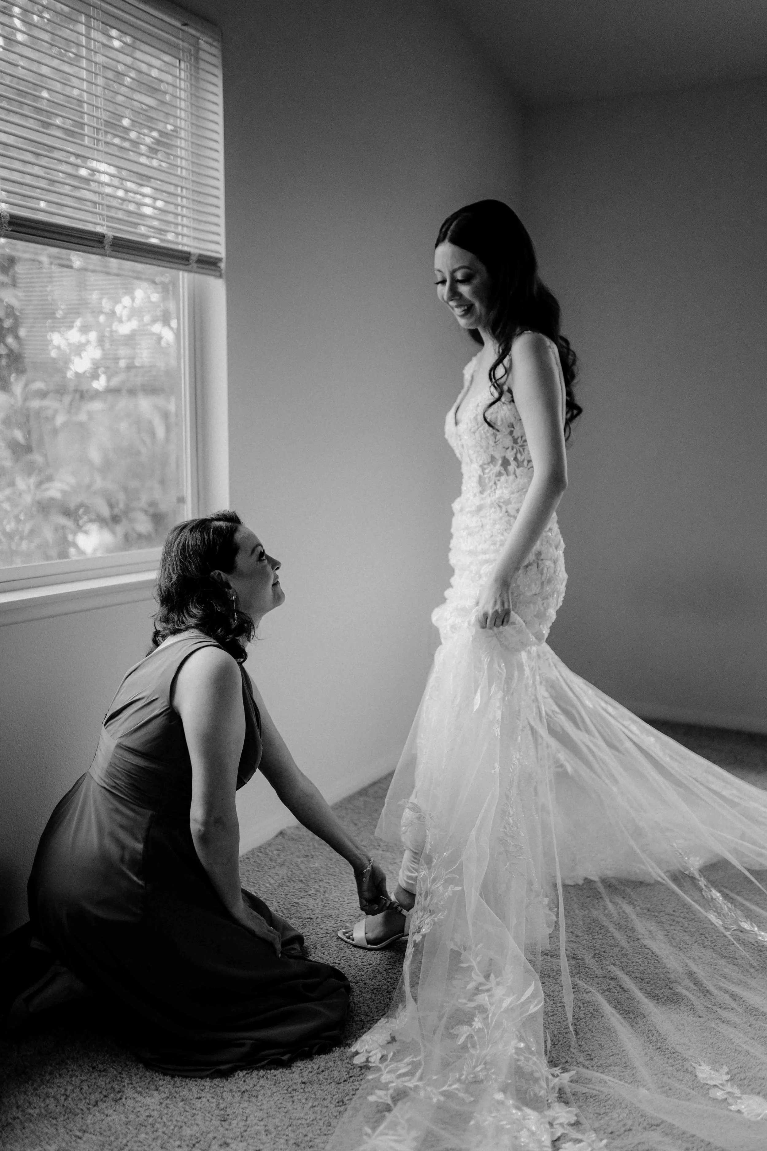A bride in a lace wedding dress standing and smiling while a woman in a sleeveless dress kneels and helps her with her shoe near a window.