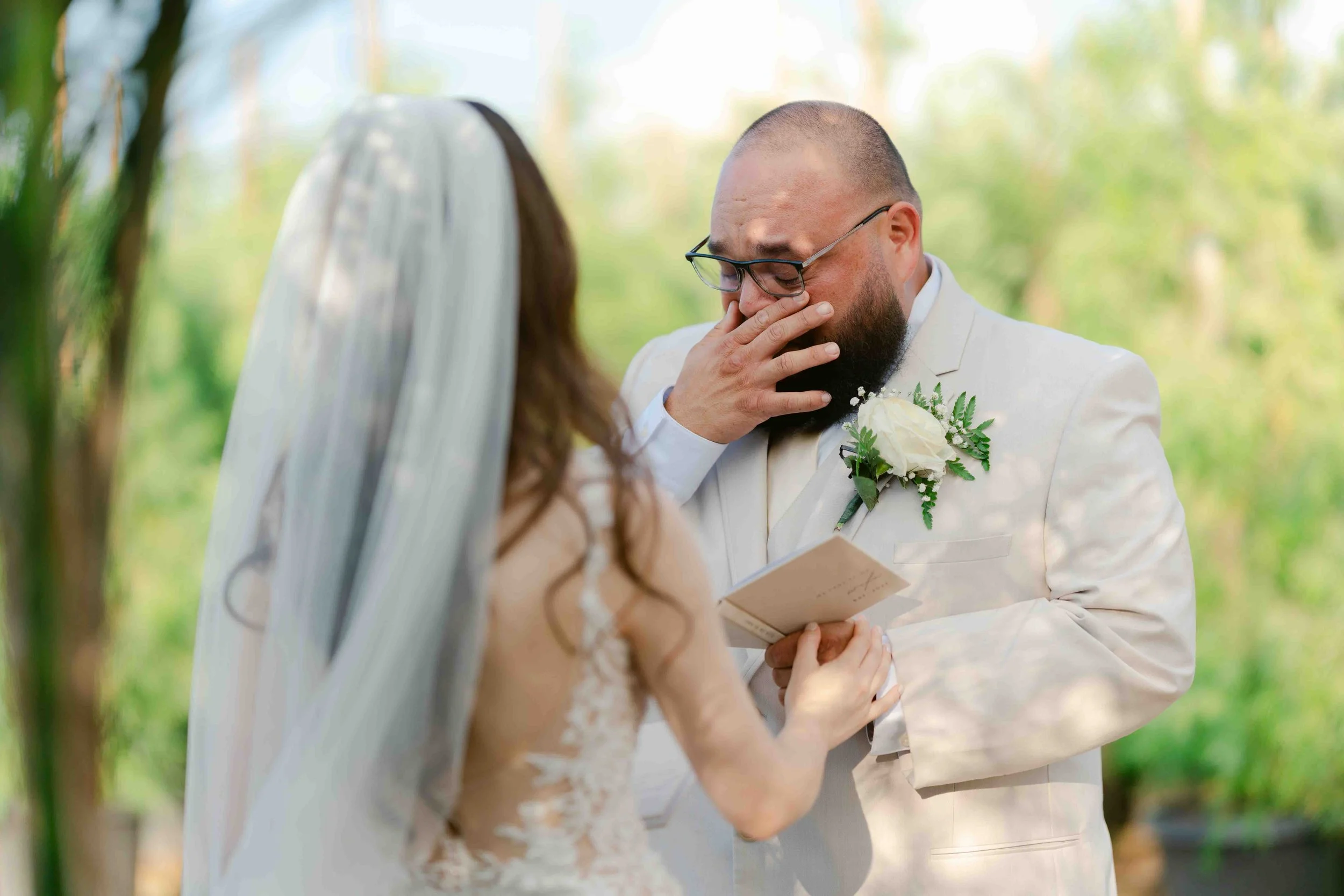 Groom in a white suit crying during a wedding ceremony outdoors, with a bride in a wedding dress and veil, holding a small book or note.