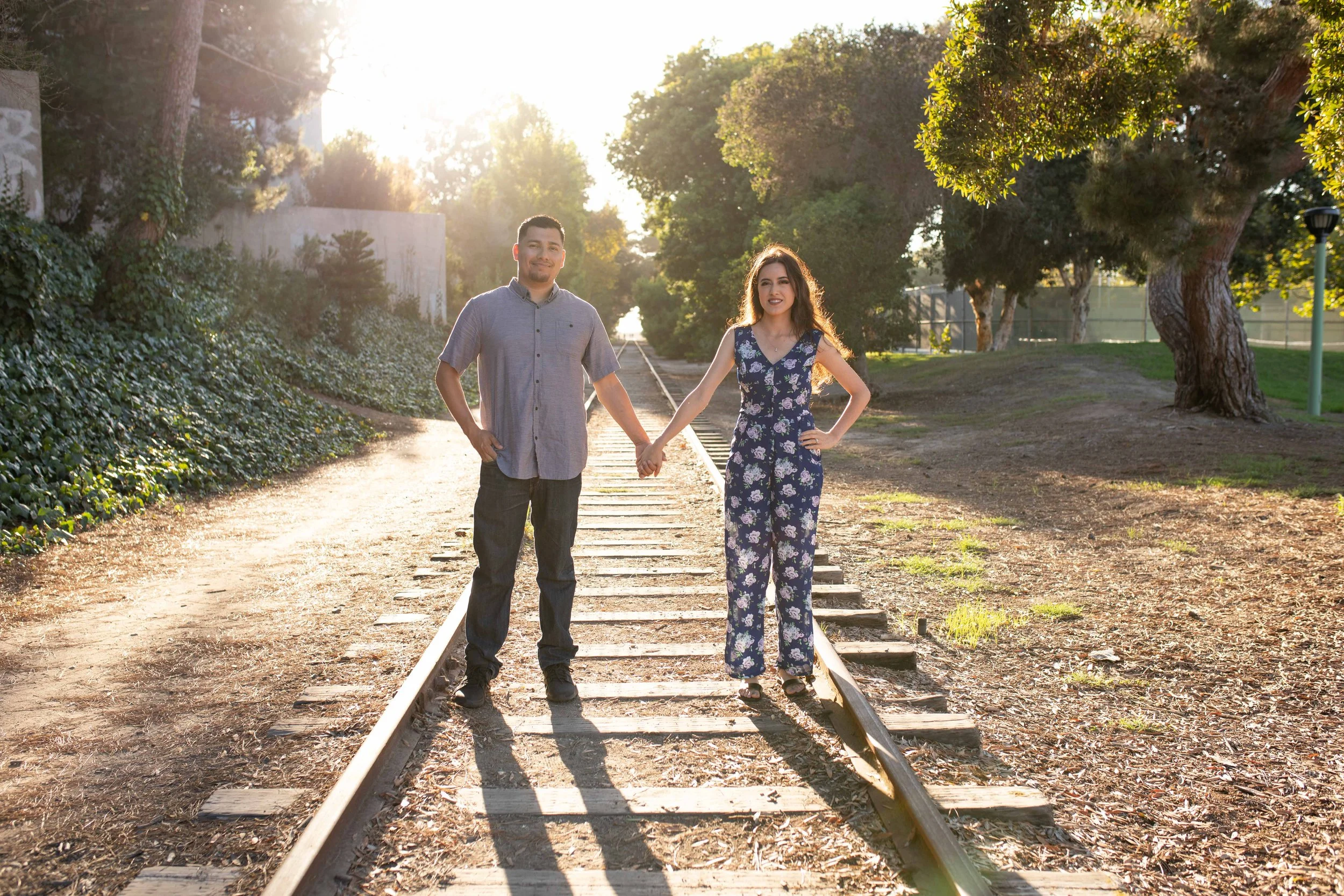 A man and woman holding hands on a railway track in a park during sunset, surrounded by trees and greenery.