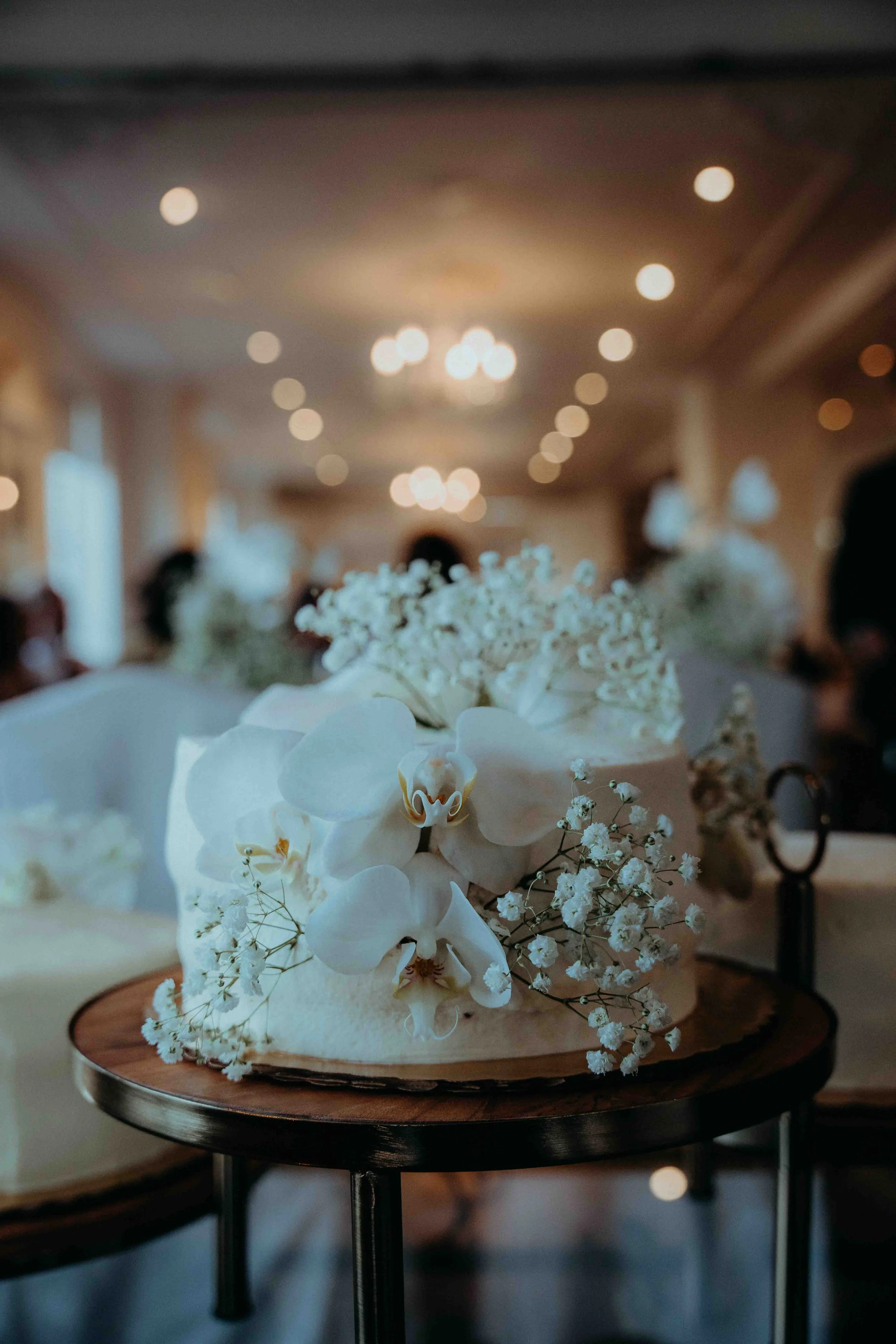 A wedding cake decorated with white orchids and baby's breath flowers, placed on a wooden stand in a banquet hall.