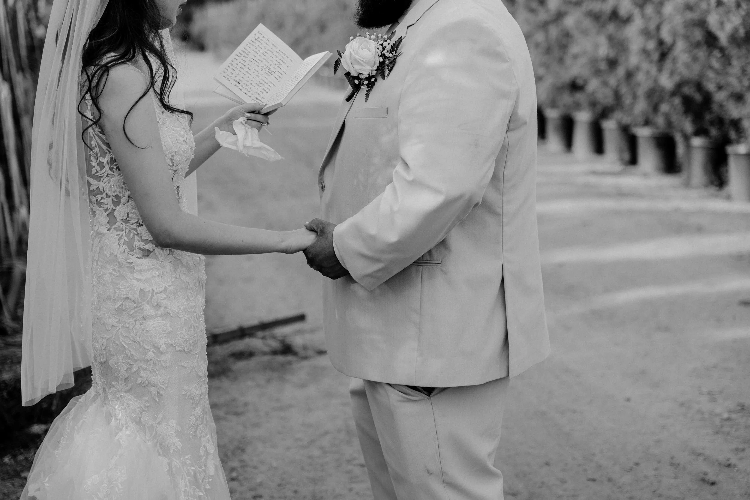 A black-and-white photo of a bride and groom holding hands during their wedding ceremony, with the bride reading vows from a paper.