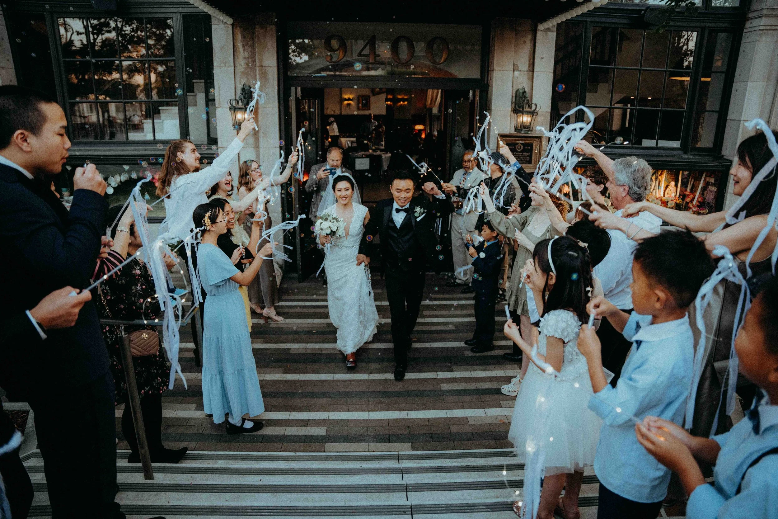 Bride and groom walking out of a building through a crowd of friends and family holding sparklers and streamers during a wedding celebration.