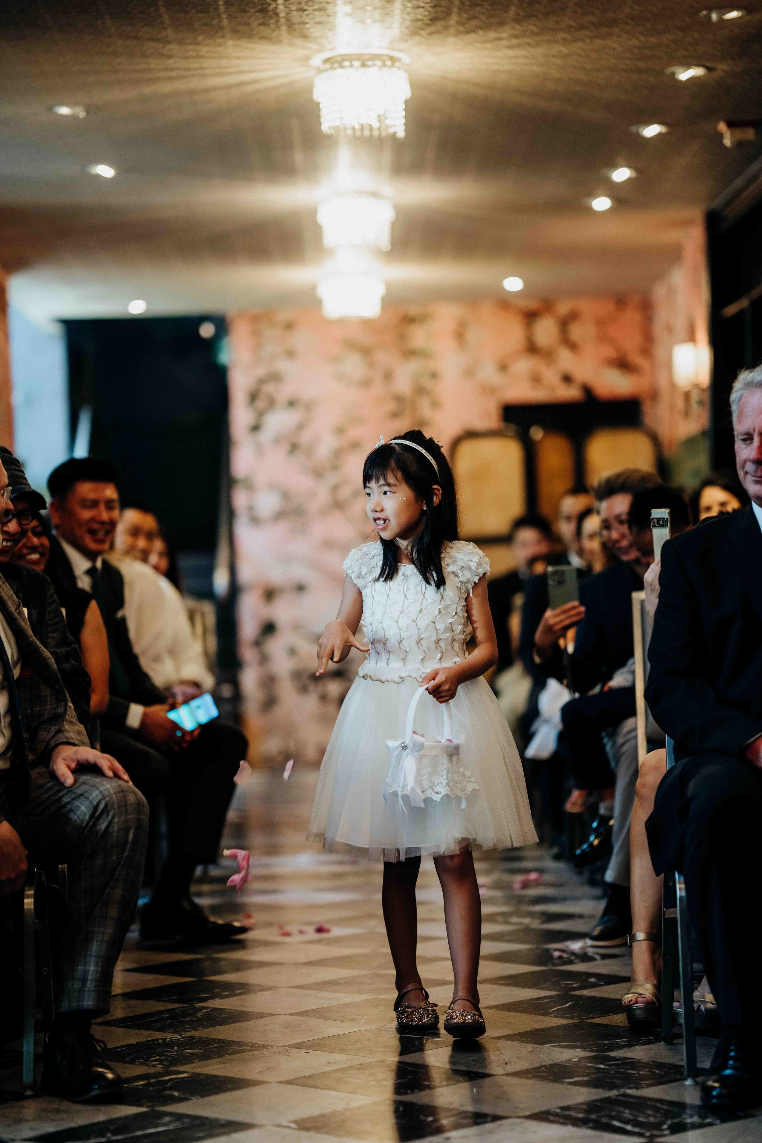 A young girl in a white dress is walking down an aisle at a wedding, holding a basket and surrounded by seated guests.