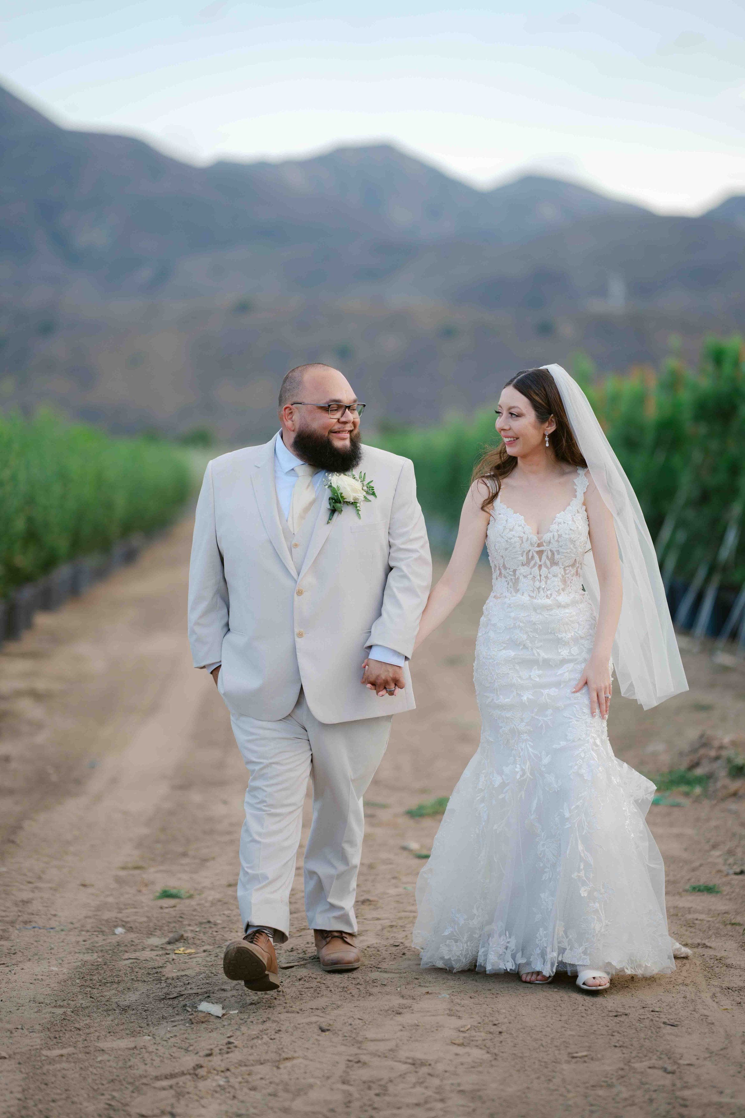 A newlywed couple walking hand in hand on a dirt path, with green plants flanking the pathway and mountainous terrain in the background, during early evening.