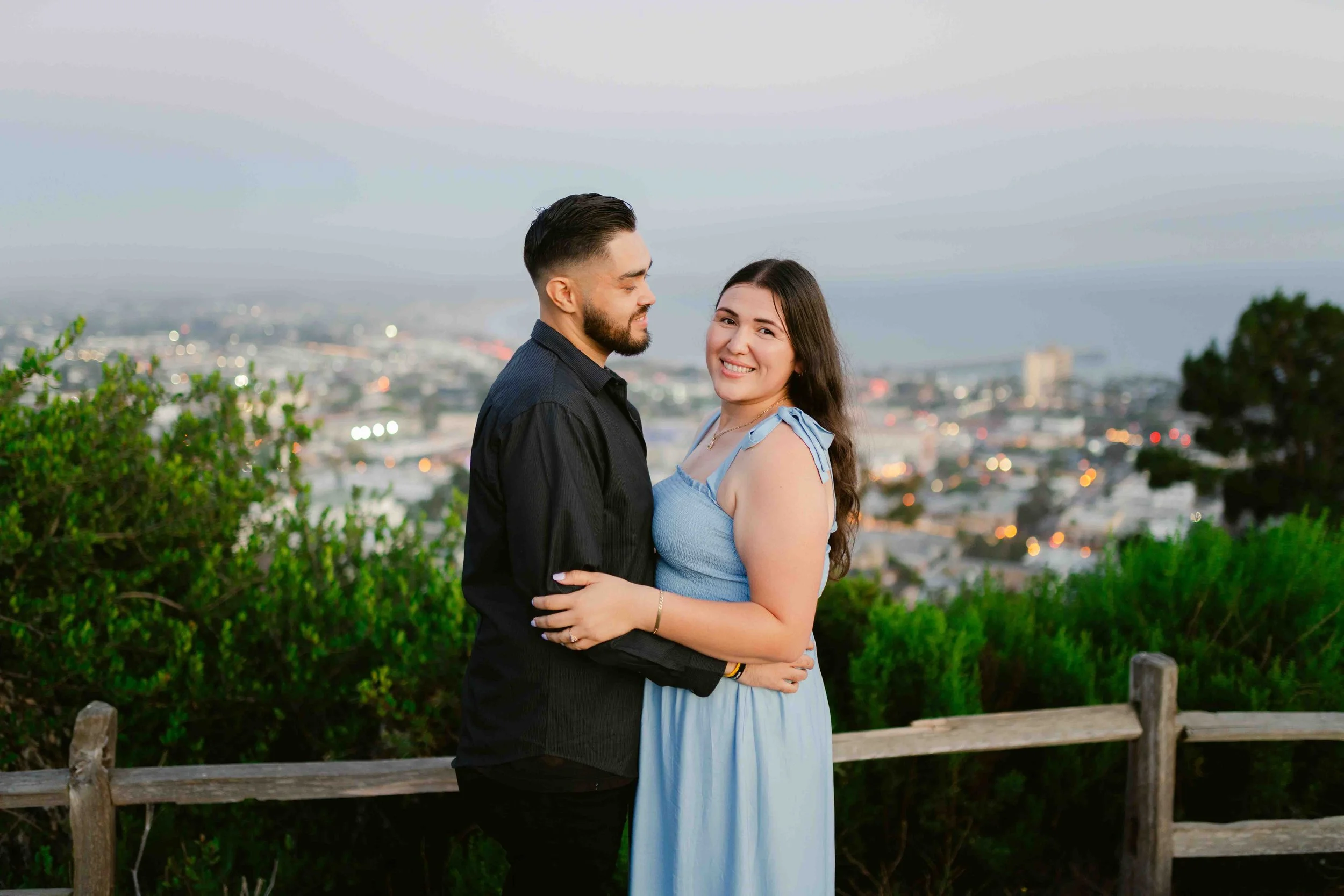 A smiling young couple hugging on an outdoor hilltop with a cityscape in the background during dusk.