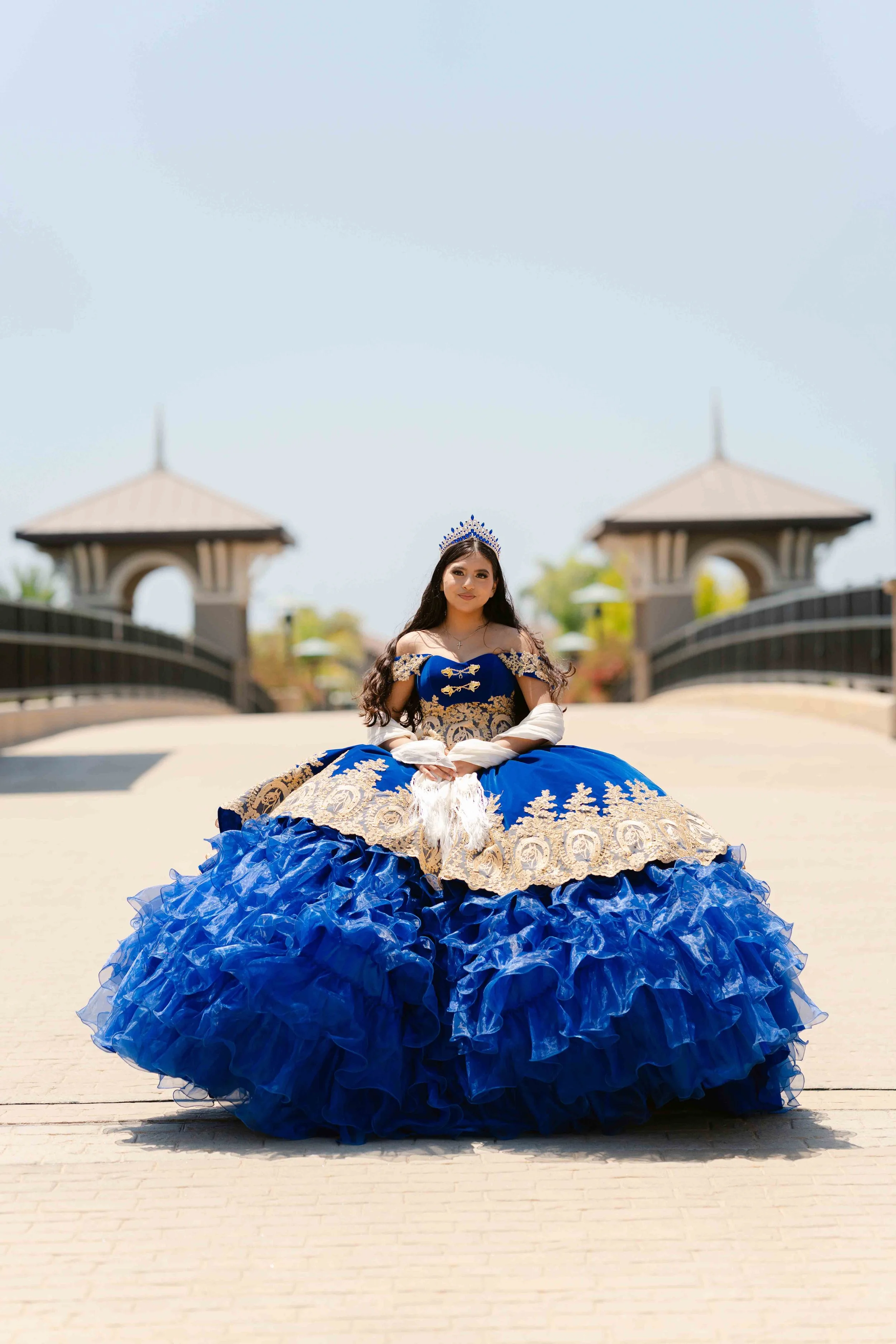 A young woman in a royal blue and gold gown with a tiara, sitting outdoors on a paved pathway with a bridge and pavilions in the background.