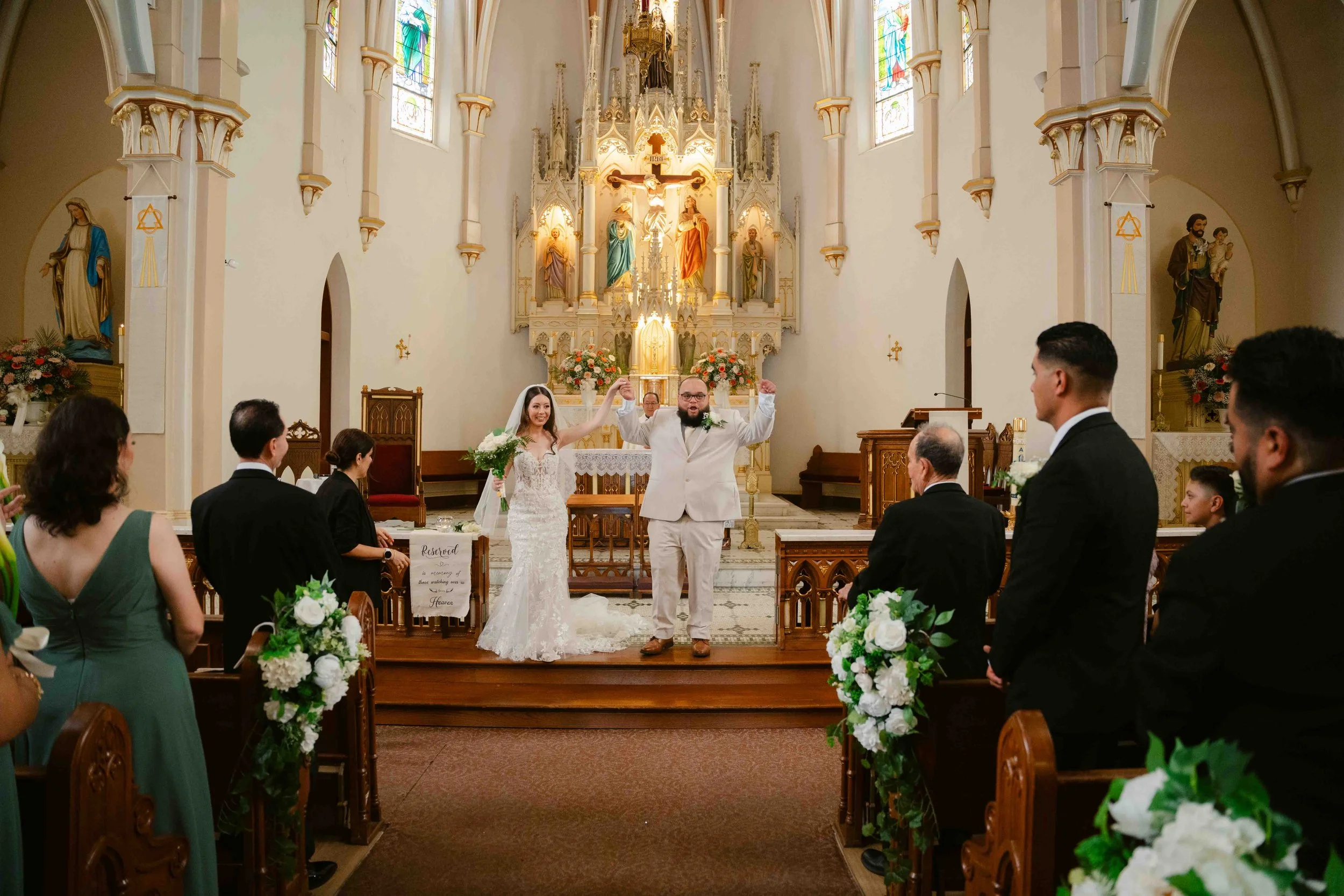 Bride and groom celebrating at their wedding ceremony inside a church, with guests watching and smiling, floral decorations, and religious statues.