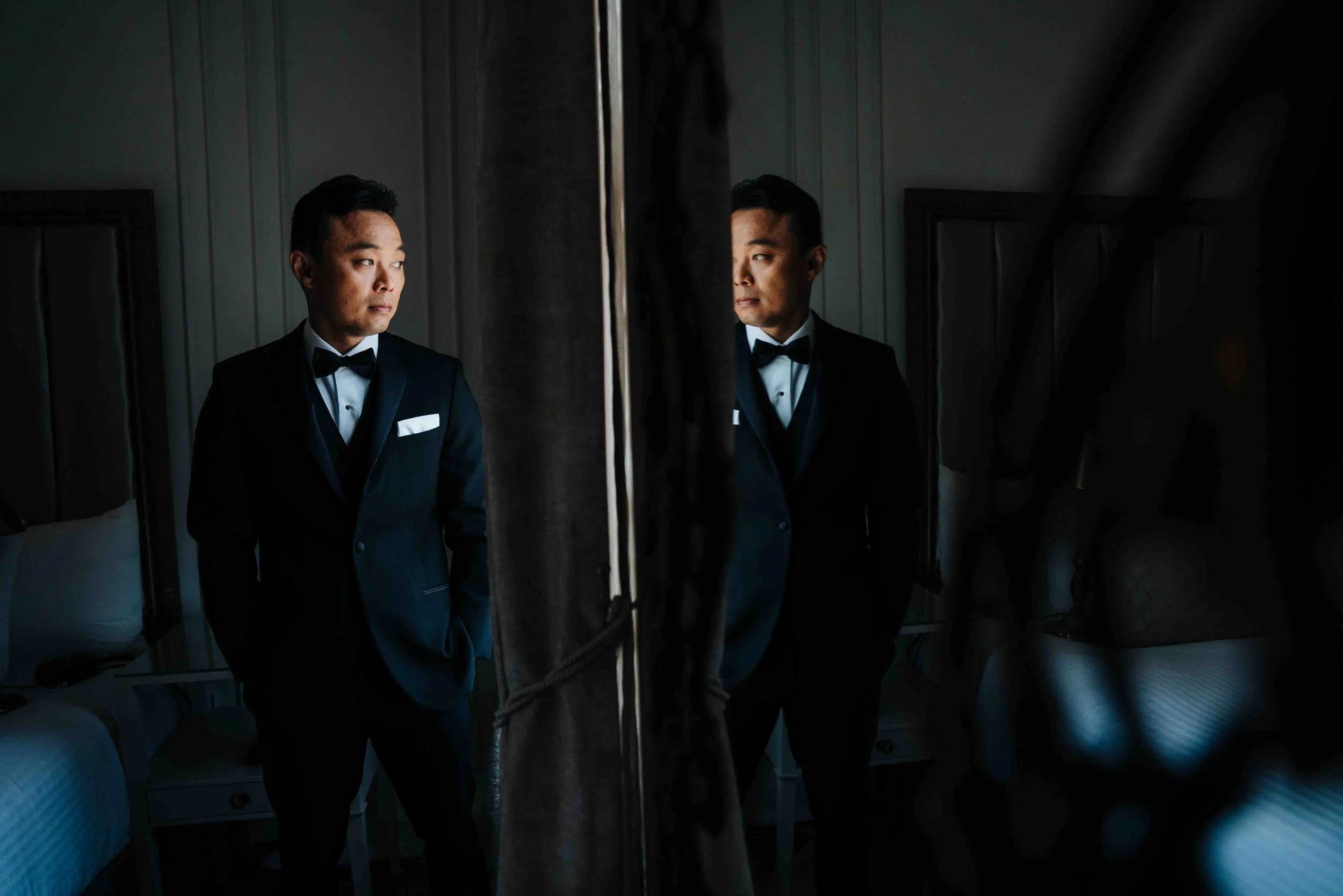 A man in a black tuxedo looking at his reflection in a mirror in a dimly lit room.