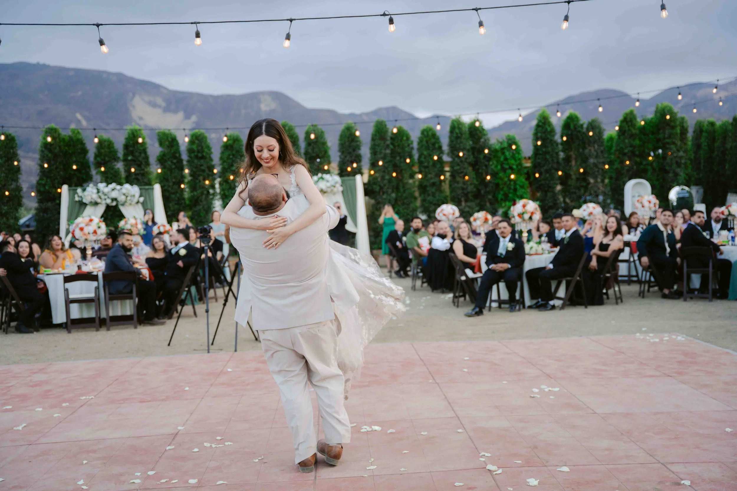 A bride and groom dancing at their wedding reception outdoors with mountains in the background, surrounded by guests seated at decorated tables under string lights.
