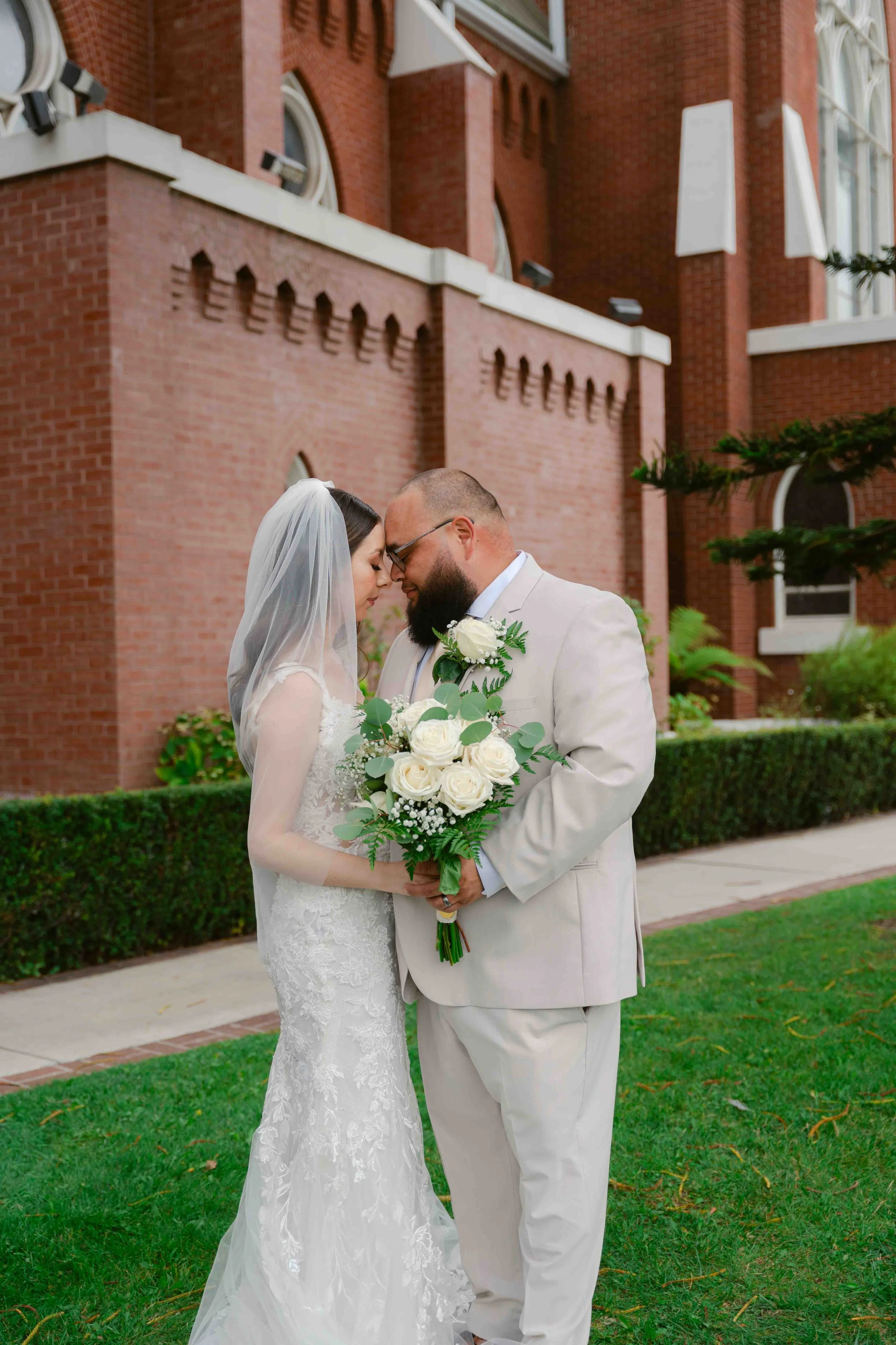 A bride and groom embrace during their wedding, with their foreheads touching and eyes closed. They are outside in front of a red brick building, with the bride holding a bouquet of white roses and greenery.
