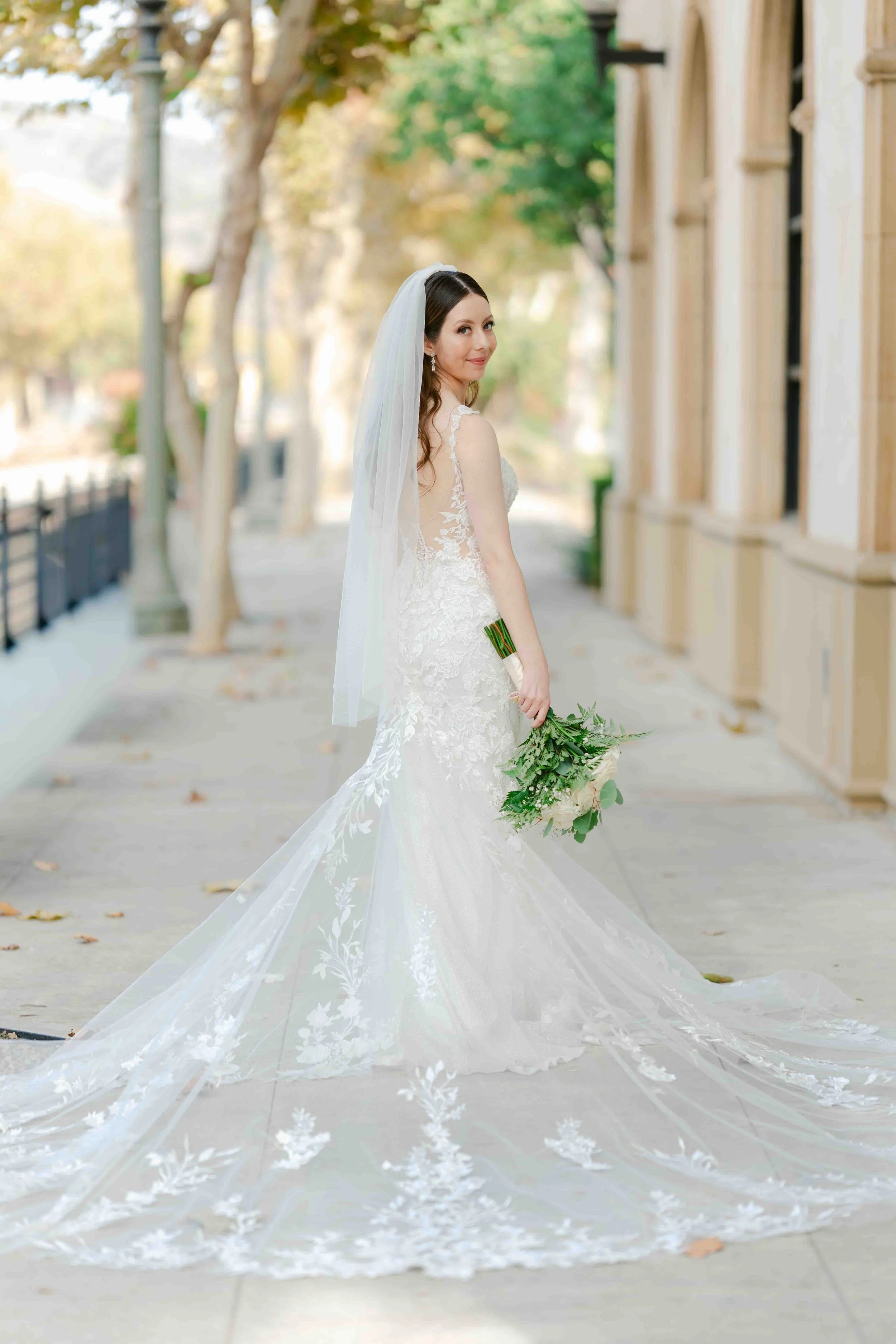 A bride in a white lace wedding gown holding a bouquet of greenery and white flowers, standing on a sidewalk with trees and buildings in the background.