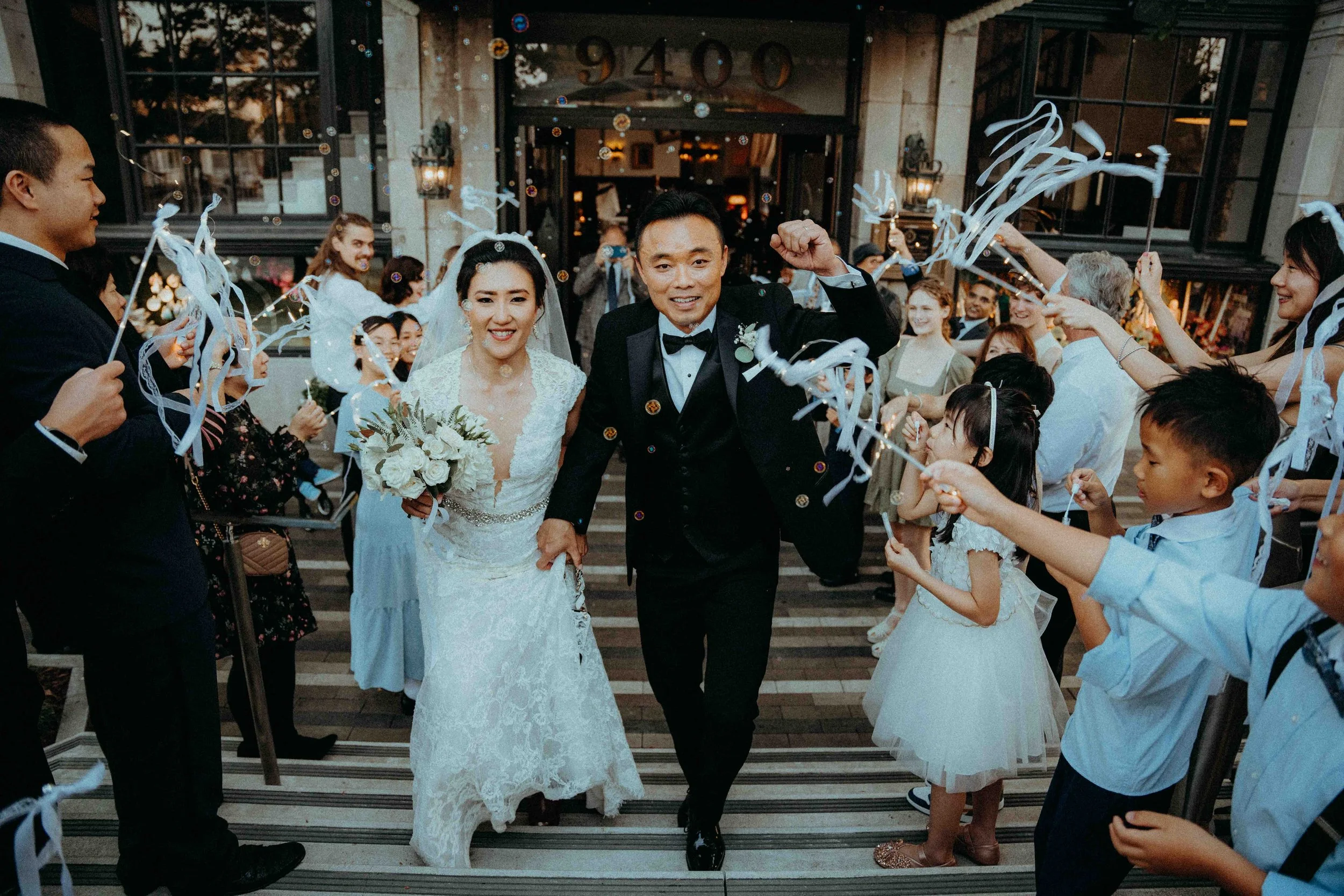 A newly married couple exiting a building, holding hands, surrounded by friends and family celebrating with streamers and bubbles outside a venue called 9400 at sunset.